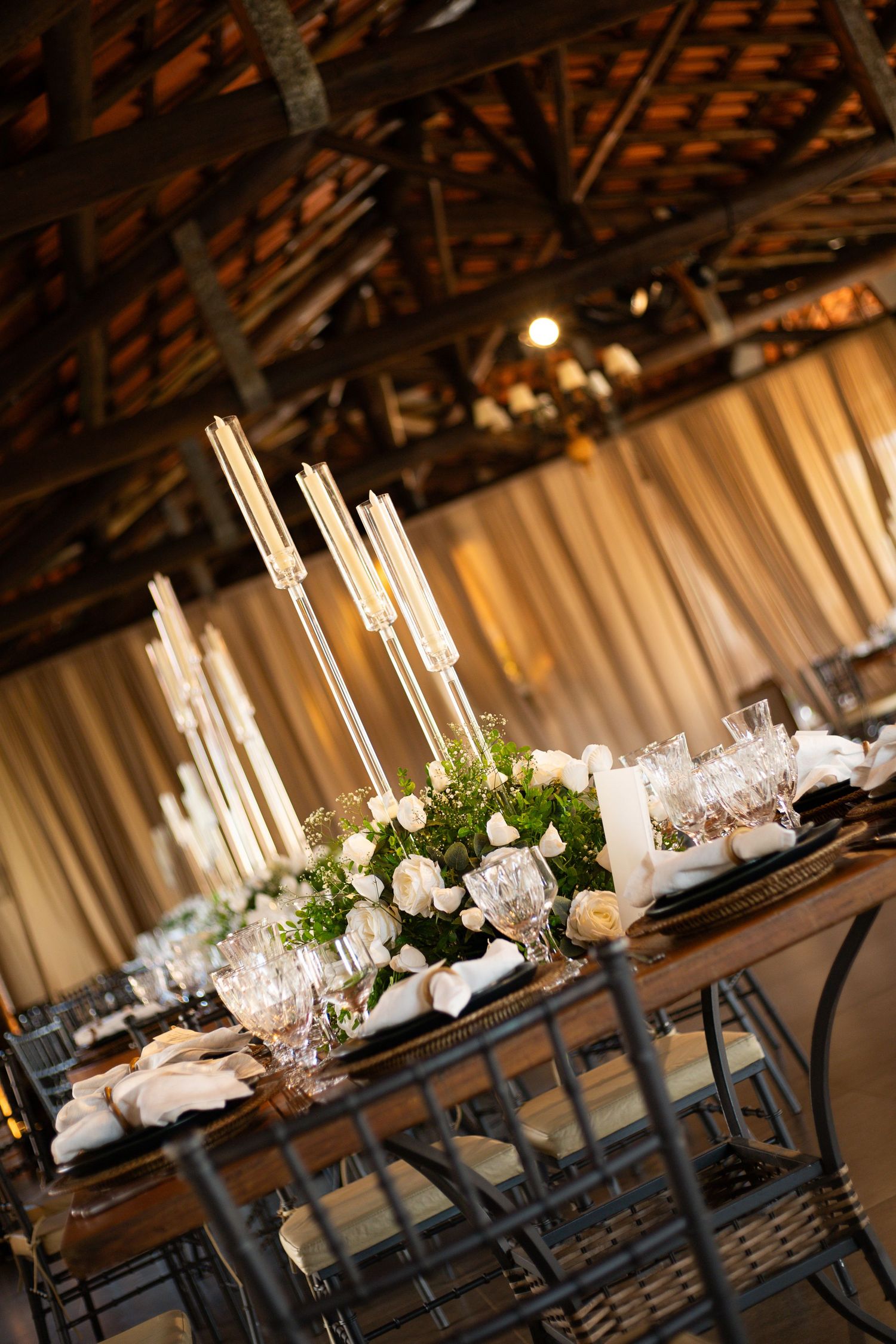 Elegant barn wedding reception table with tall candelabras, white roses, crystal glassware, and draped fabric backdrop.
