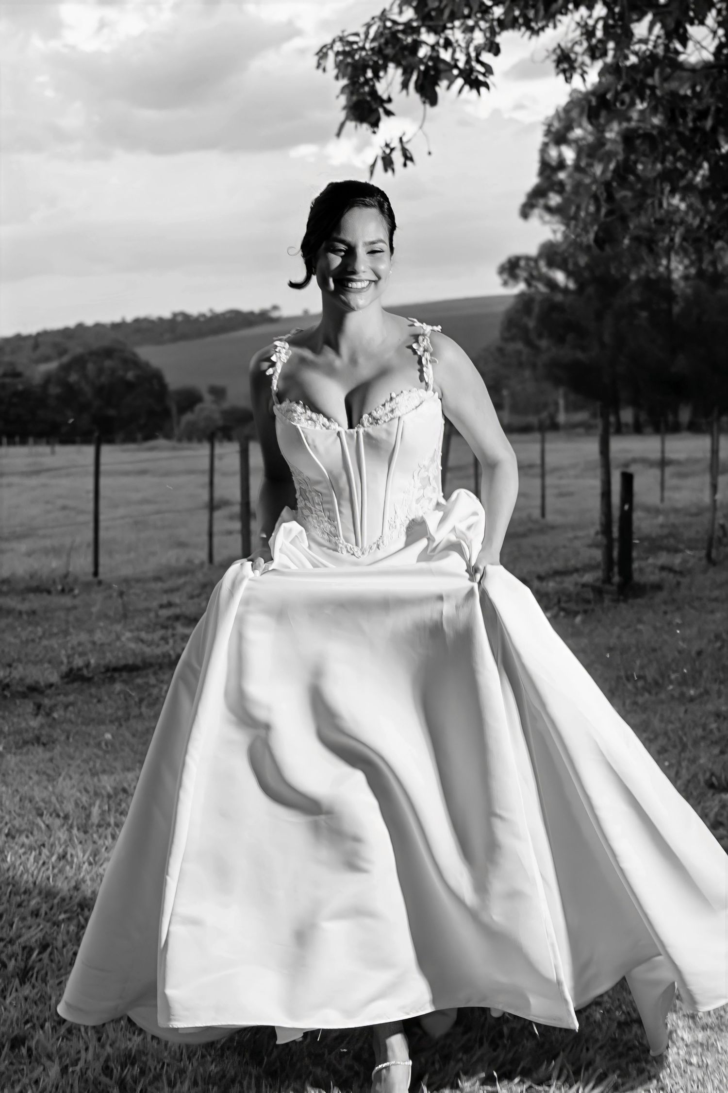 Smiling bride in a white corset wedding gown holds her skirt in a black and white outdoor photo.
