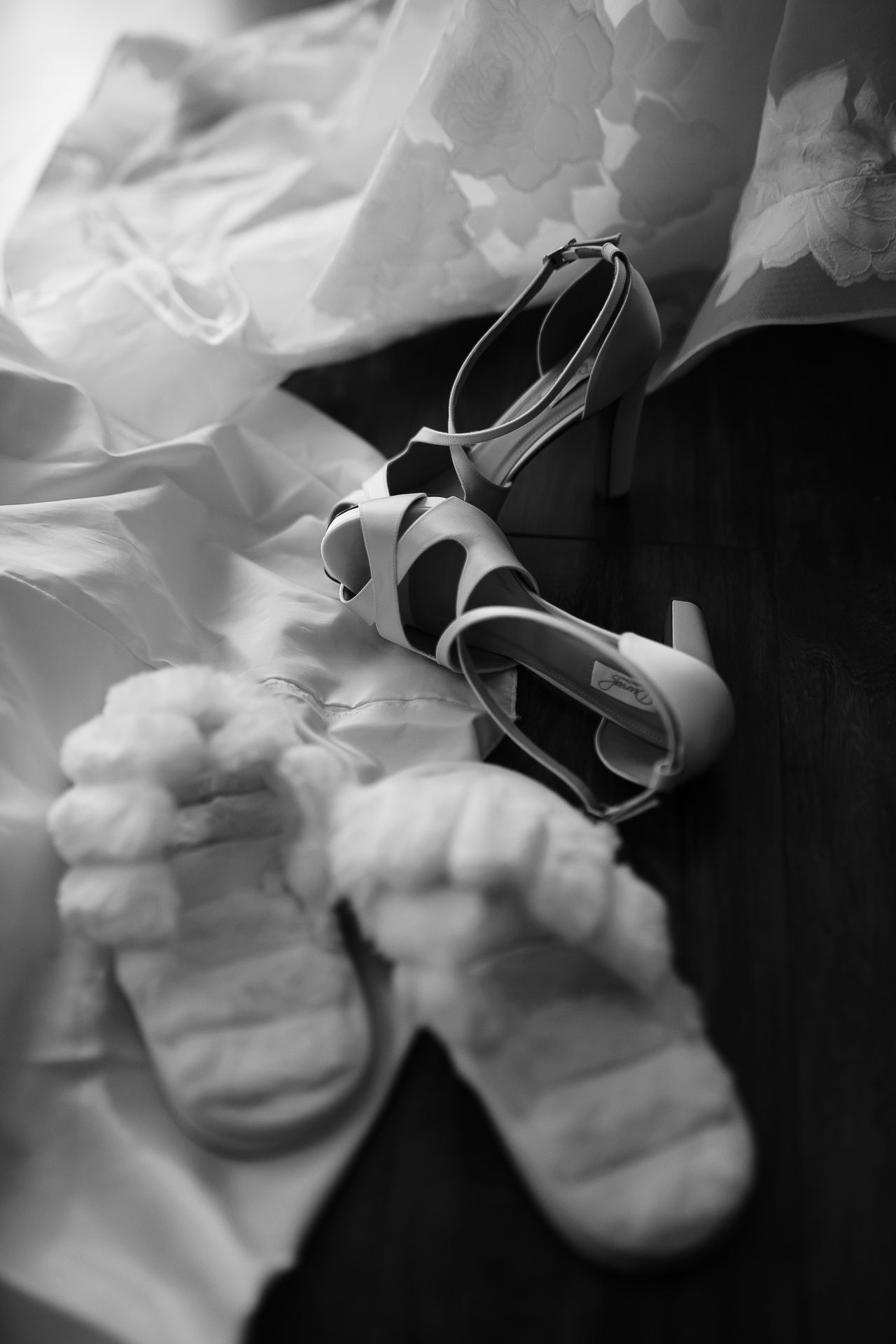Black and white photo of bridal heels and lace socks beside a wedding dress on a wooden floor.