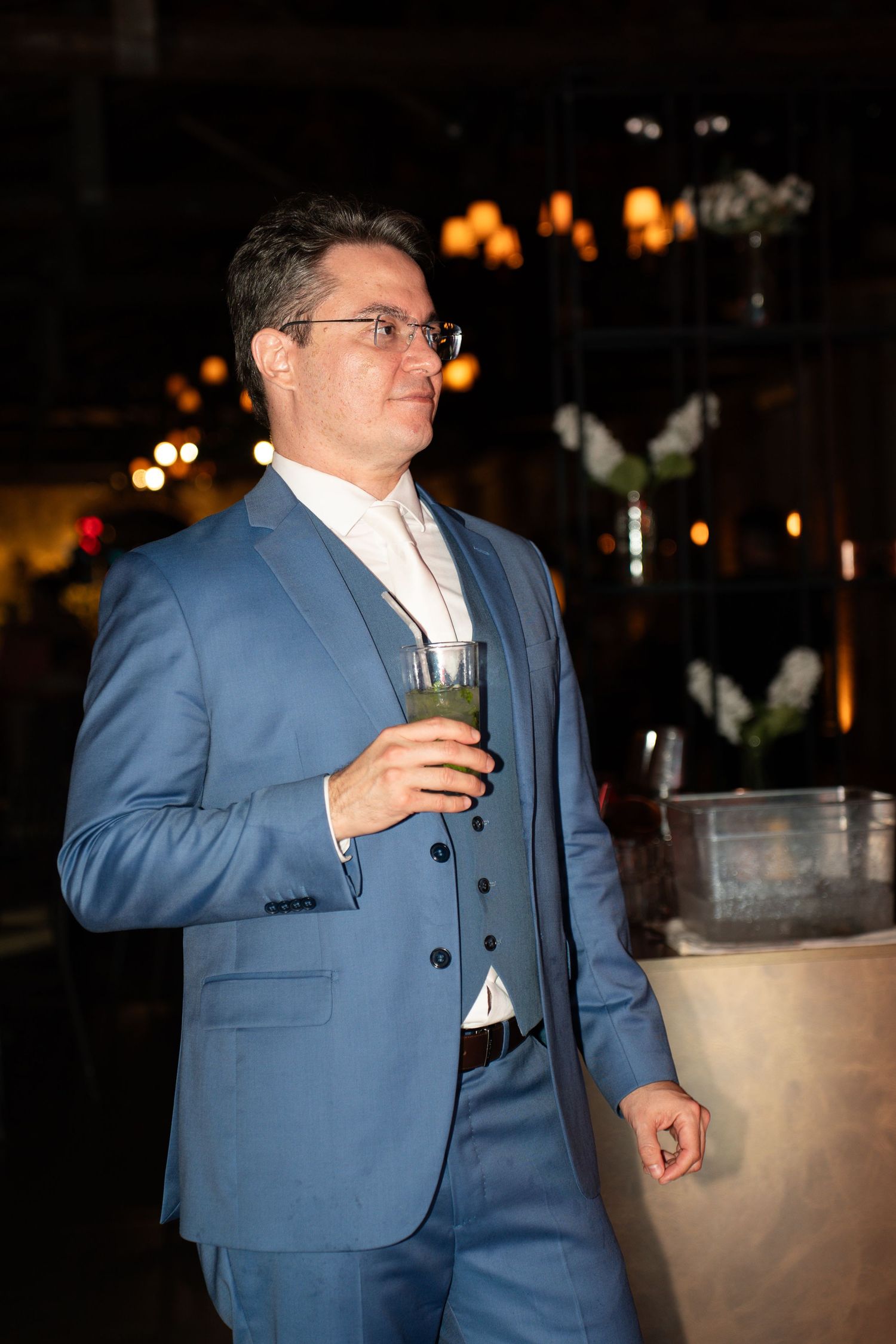 Elegant man in blue suit holding cocktail glass at upscale evening event with warm ambient lighting.