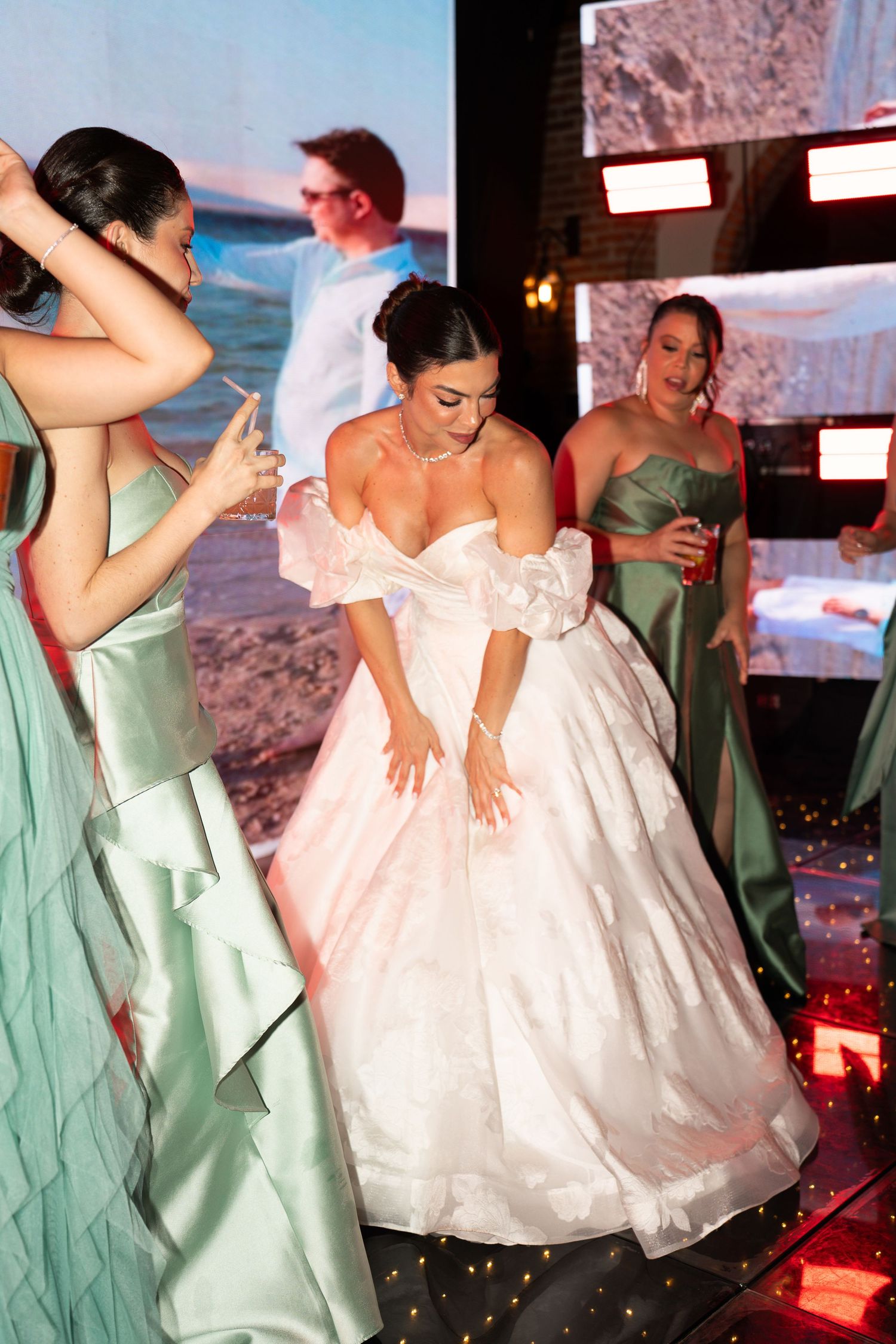 Bride in white ball gown dancing with bridesmaids in sage green dresses at elegant wedding reception.