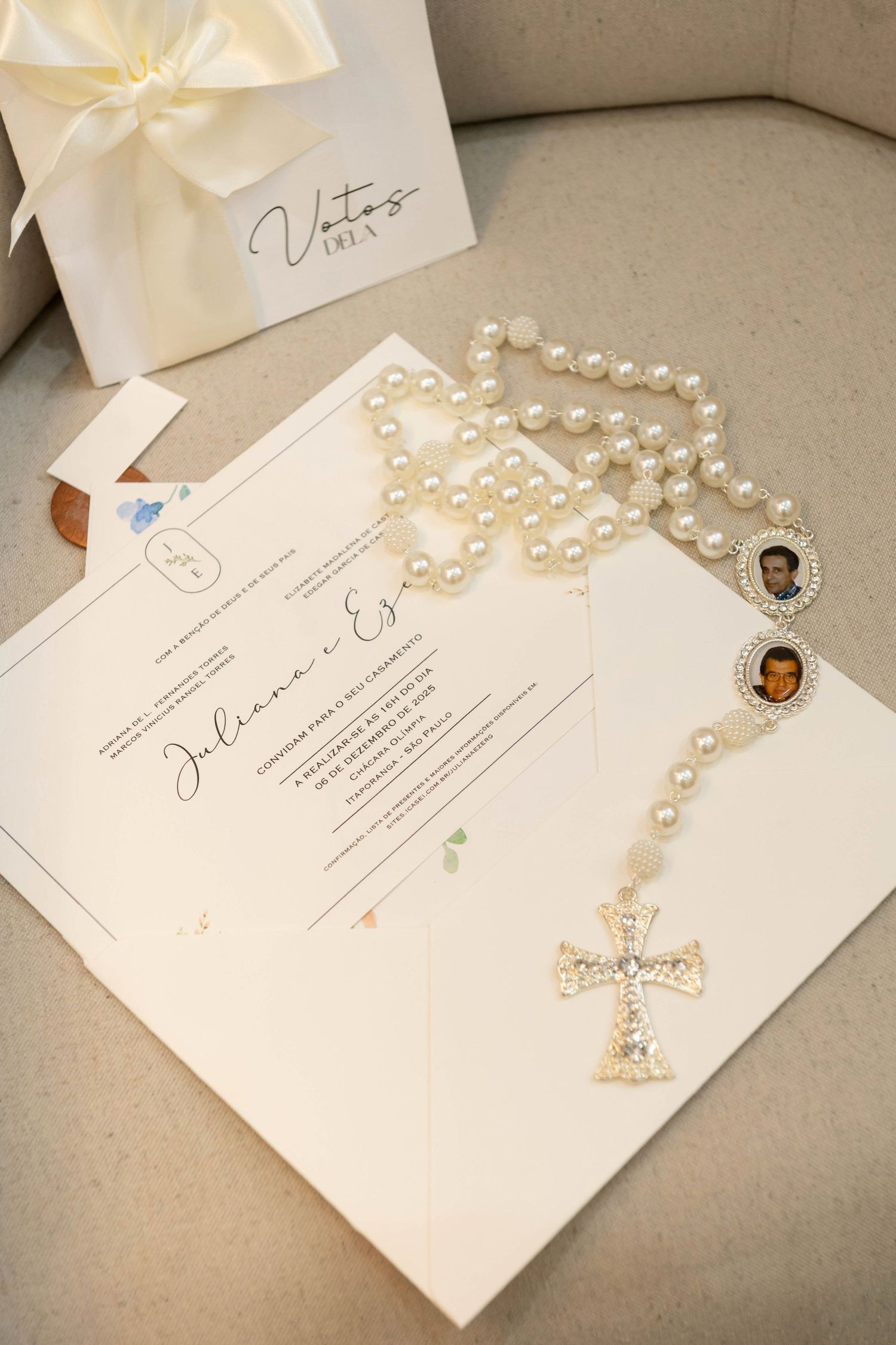 Pearl rosary with gold cross and photo charms rests on elegant wedding invitations beside a white gift box.