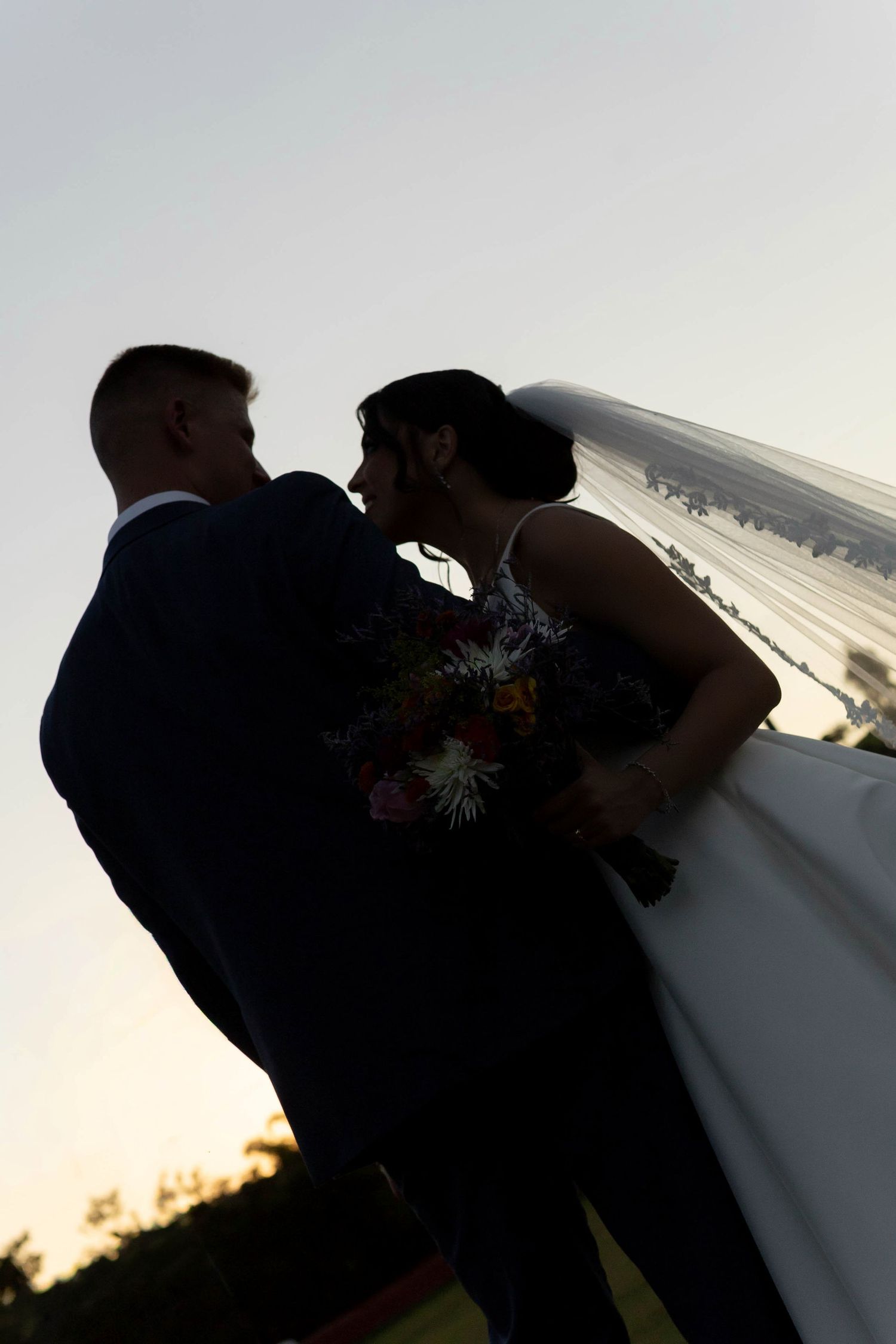 Silhouette of bride and groom sharing a romantic moment at sunset, bride holding colorful bouquet with veil flowing.