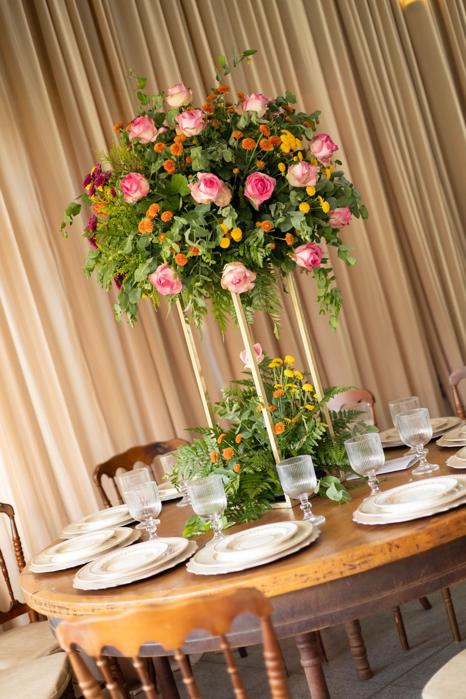 Elegant wedding table setting with tall floral centerpiece featuring pink roses and greenery on a wooden table.