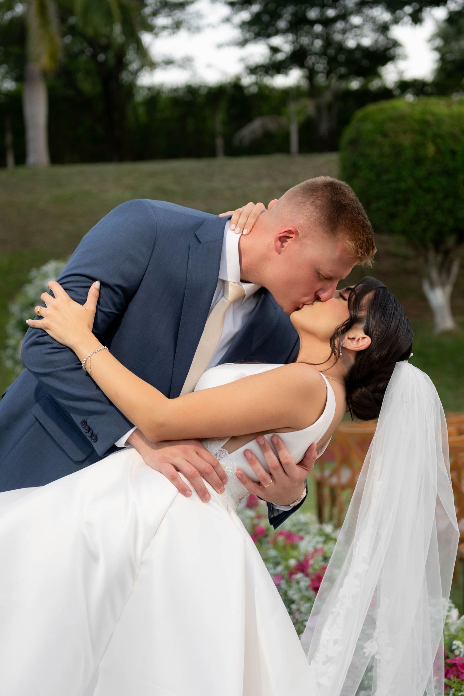 Bride and groom share a romantic dip kiss outdoors, with lush greenery in the background at their wedding.