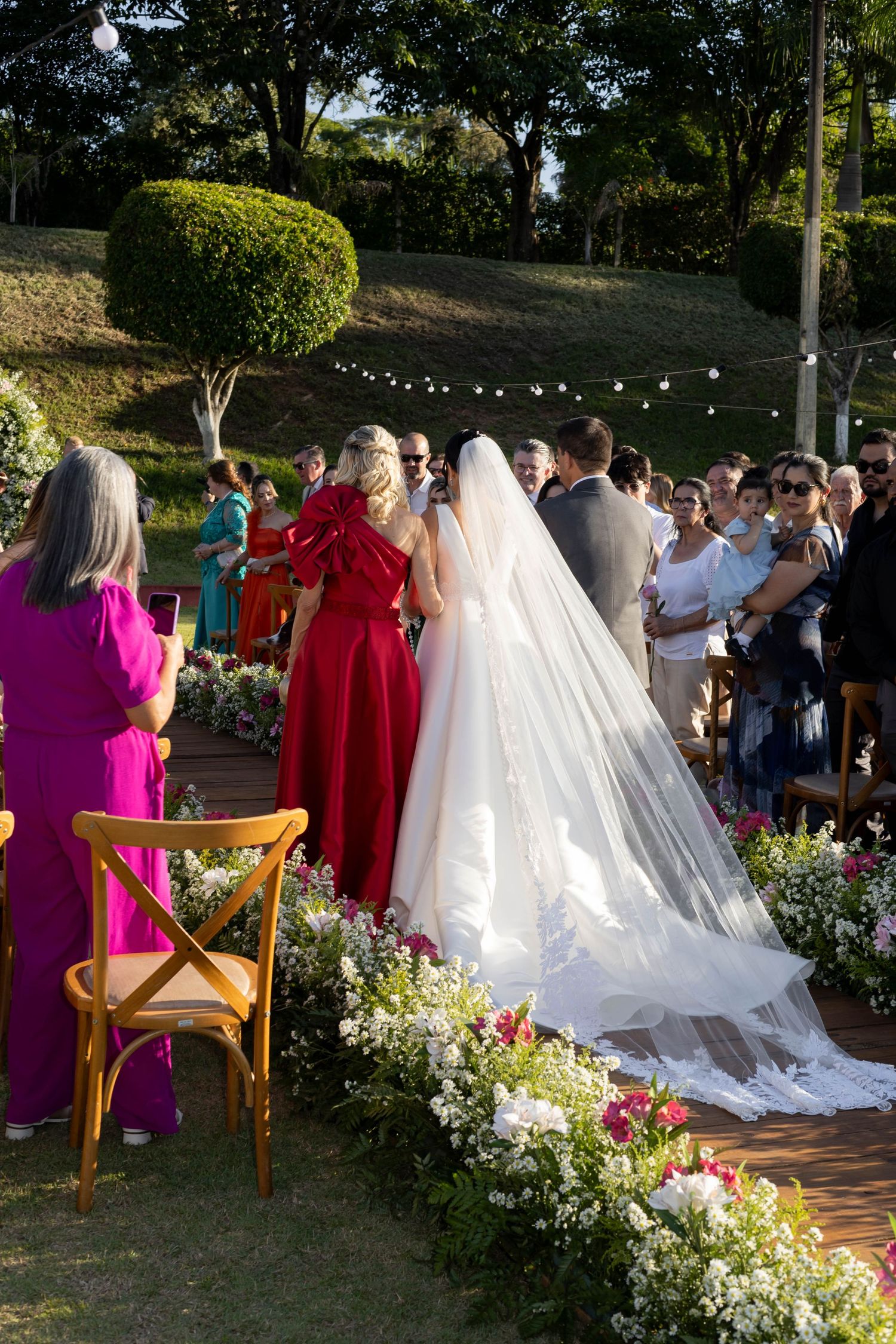 Outdoor wedding ceremony with bride in white gown and veil, guests in colorful attire, surrounded by floral arrangements and string lights.