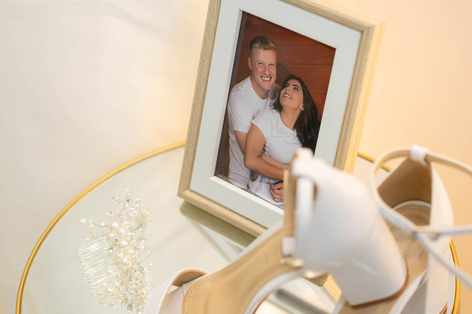 White bridal heels and crystal jewelry displayed on a glass table beside a framed photo of a smiling couple embracing.