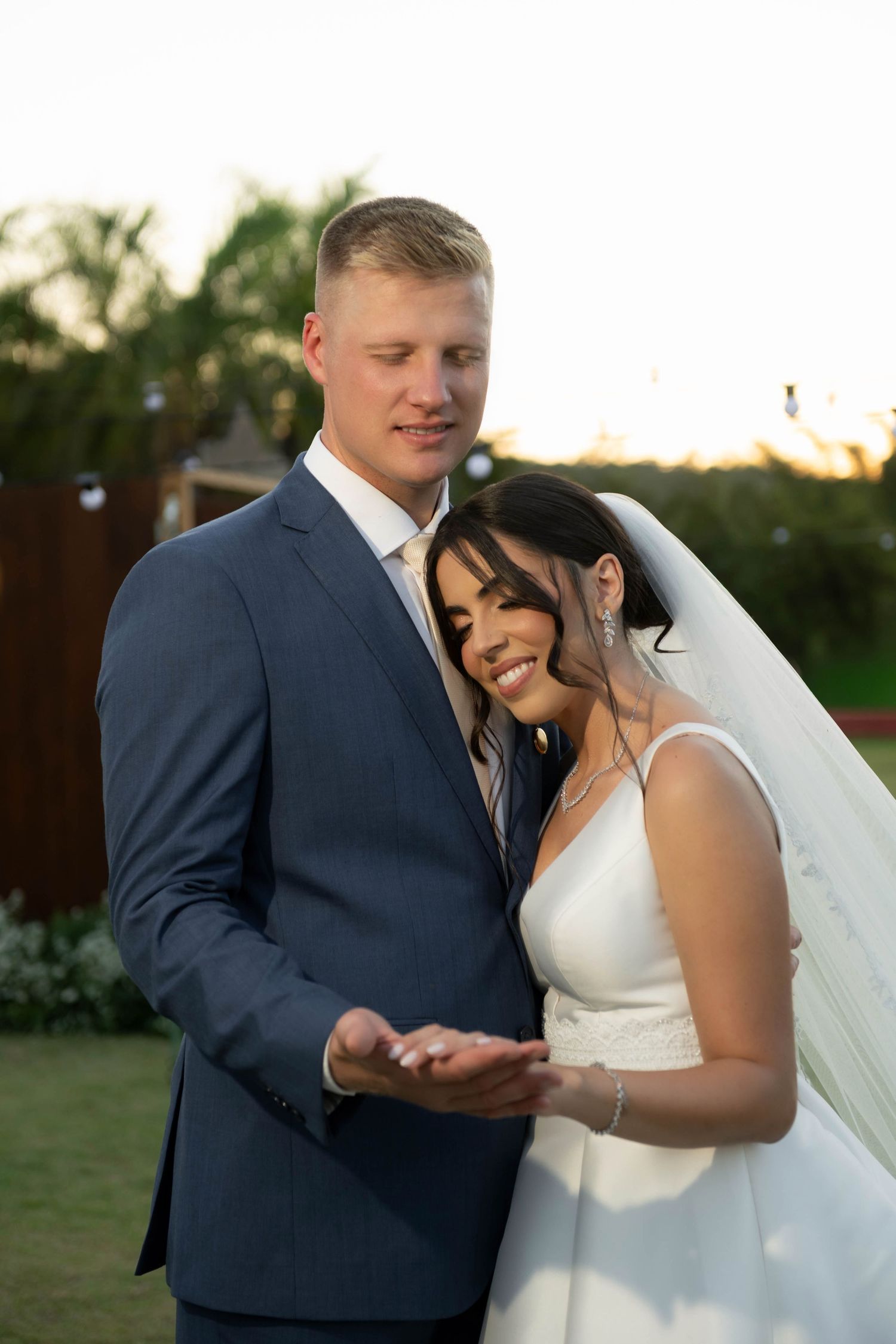 Bride and groom exchanging rings outdoors at golden hour, bride smiling in white gown and veil, groom in navy suit.