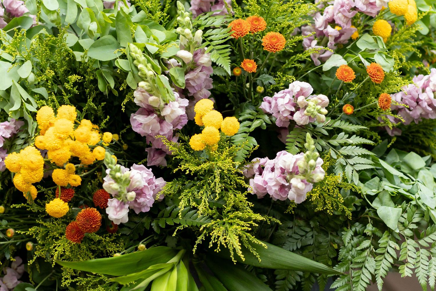 Colorful floral arrangement with yellow, pink, and orange blooms surrounded by lush green ferns and foliage.
