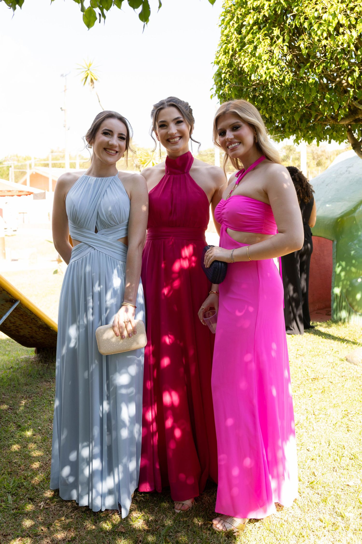 Three women in elegant formal gowns, silver, magenta, and pink, posing outdoors at a garden event.