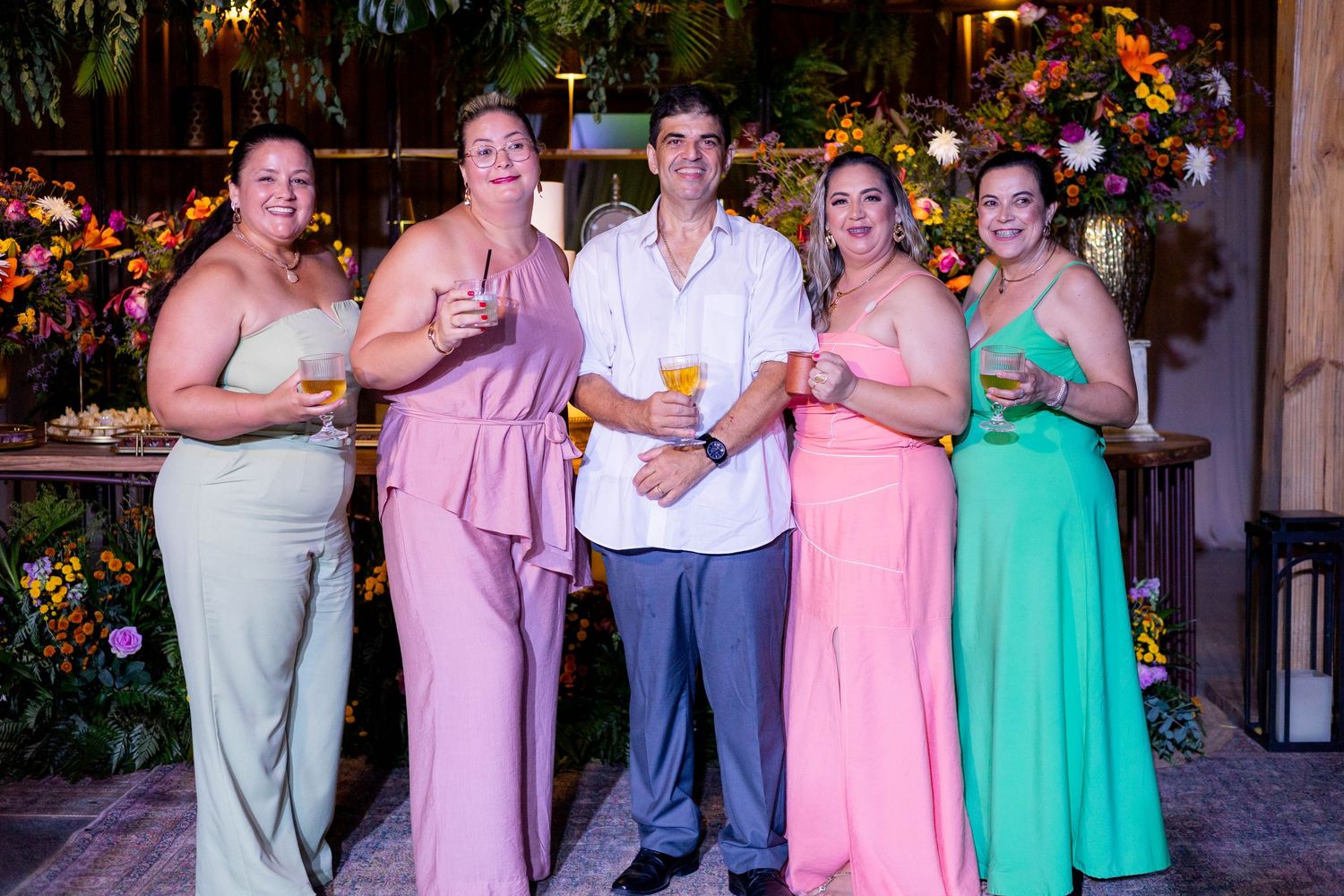 Five elegantly dressed guests pose together at a floral-decorated event, holding cocktails in colorful formal attire.