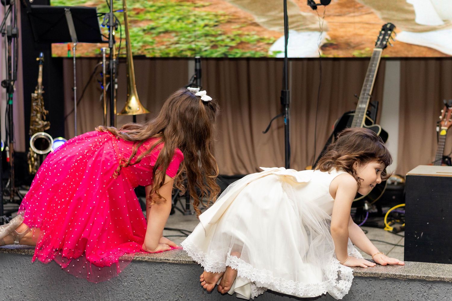 Two young girls in formal dresses crawling on stage near musical instruments at an outdoor event.