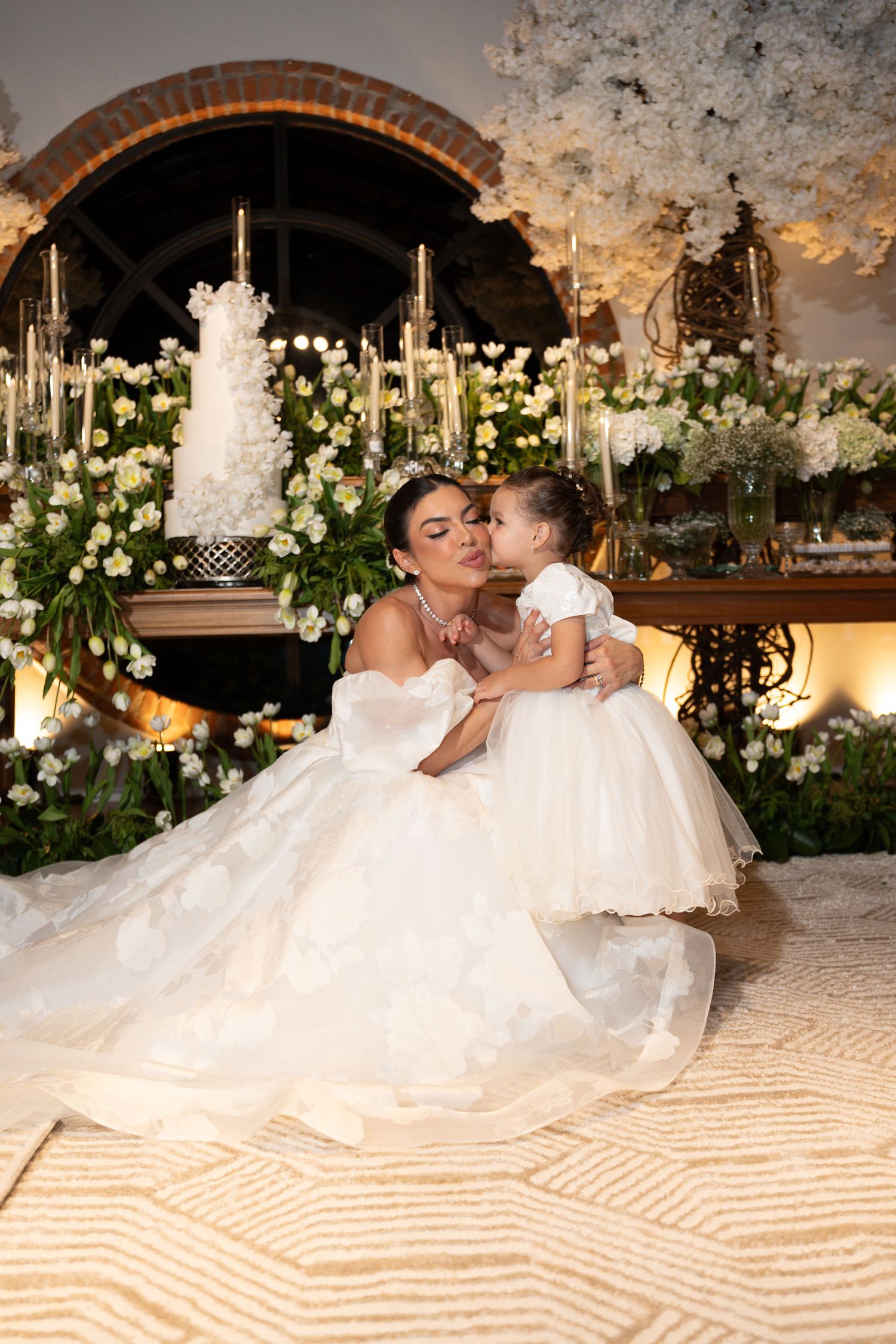 Bride in a ball gown kisses a flower girl in white dress, surrounded by floral arrangements and wedding cake.