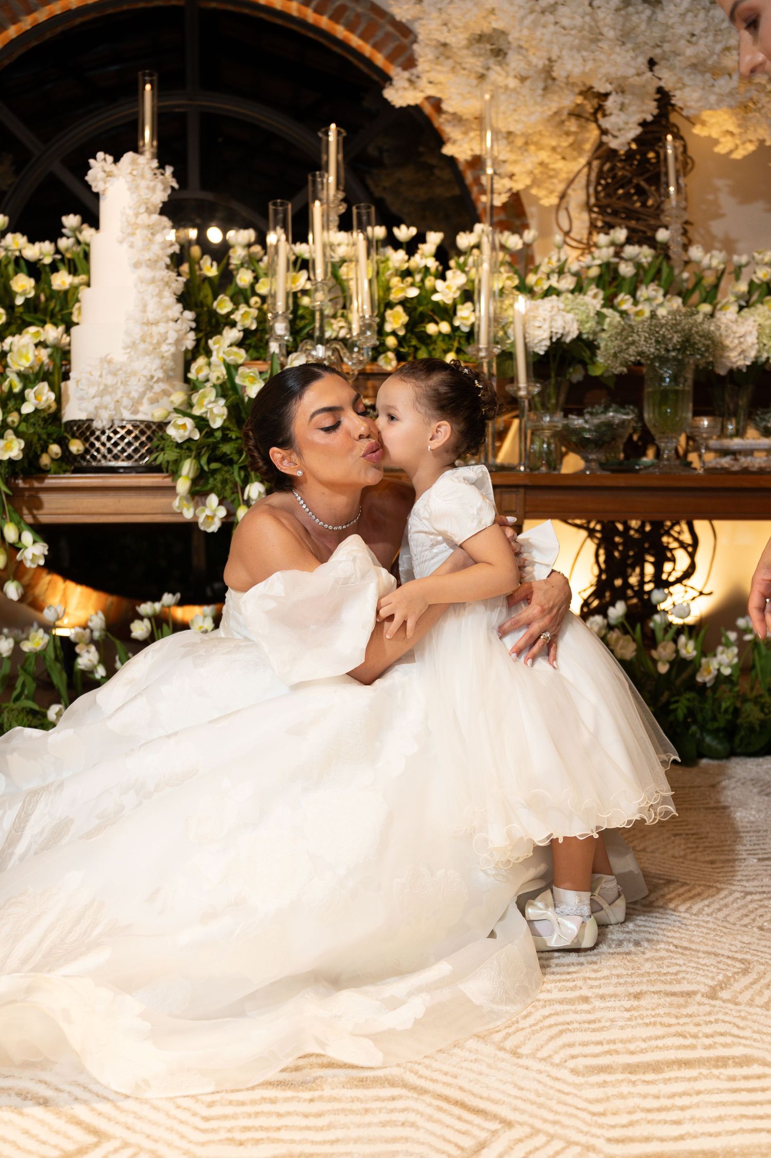 Bride in white gown kisses young flower girl in white dress at elegant floral wedding reception.