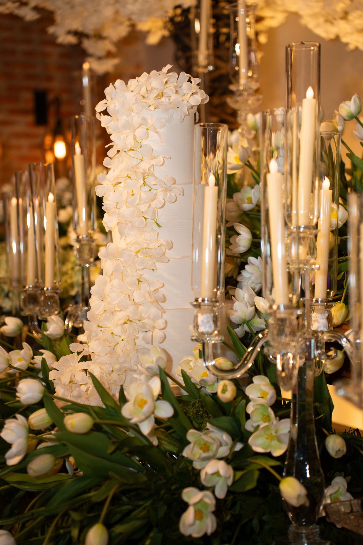 Elegant white wedding cake adorned with orchids surrounded by candles and white tulips on a decorated reception table.