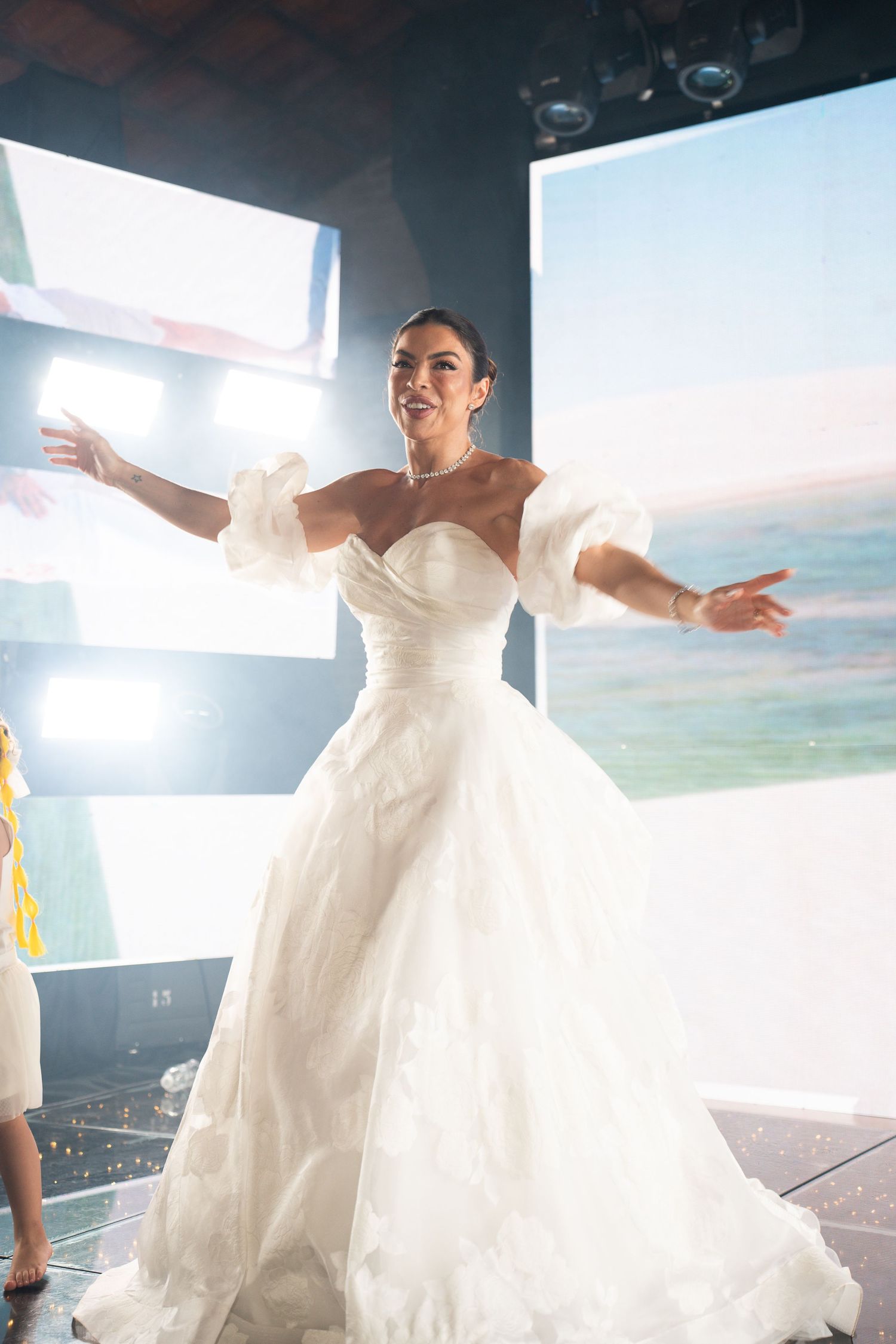 Woman in elegant white off-shoulder ball gown wedding dress posing gracefully on a stage with arms outstretched.