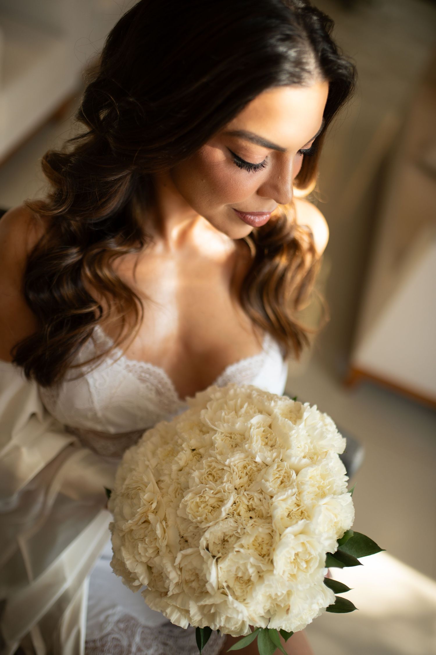 Bride in white lace gown holding large round bouquet of cream carnations, looking down with soft smile.