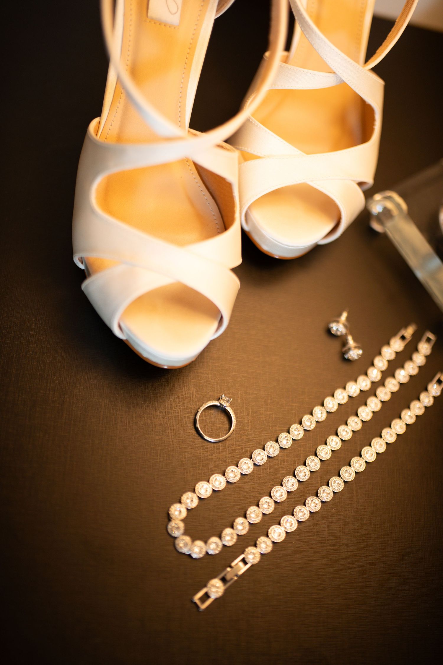 White bridal heels displayed with diamond bracelet, engagement ring, and earrings on dark surface.