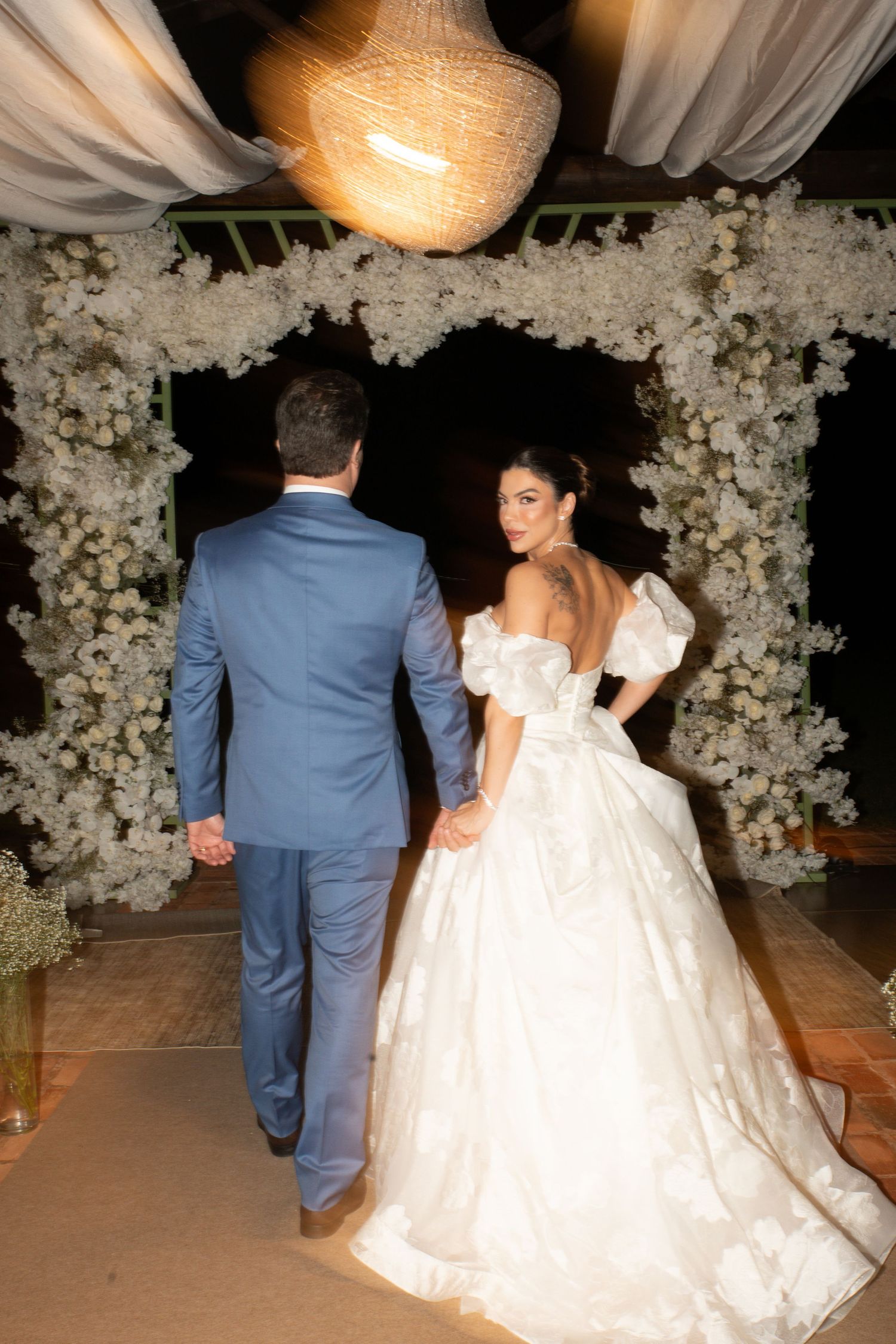 Bride in off-shoulder ball gown and groom in blue suit walk hand-in-hand under a white floral arch at night.