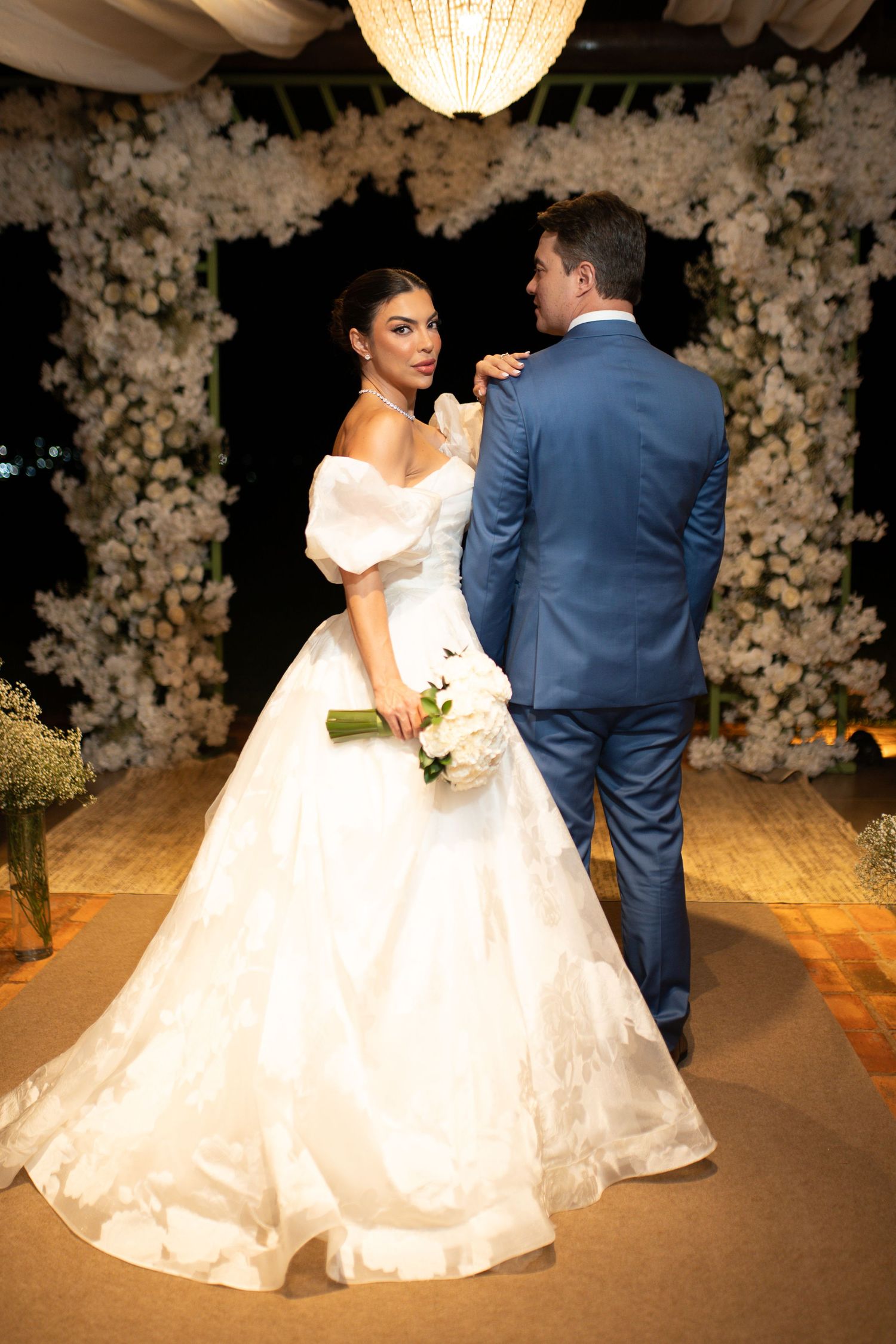 Bride in white off-shoulder ball gown holding bouquet stands with groom in blue suit before floral arch.