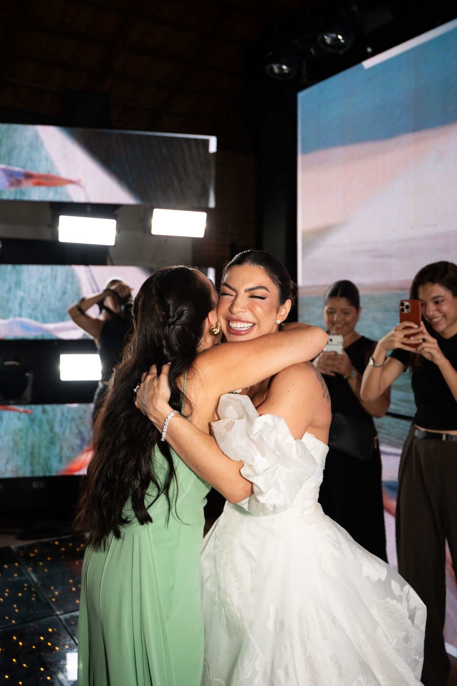Bride in white strapless gown hugging a guest in green dress at a wedding reception with guests photographing.
