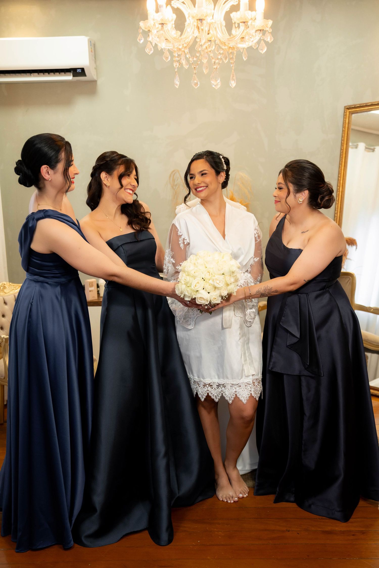 Bride in white robe holds bouquet surrounded by three bridesmaids in navy blue gowns in elegant room.