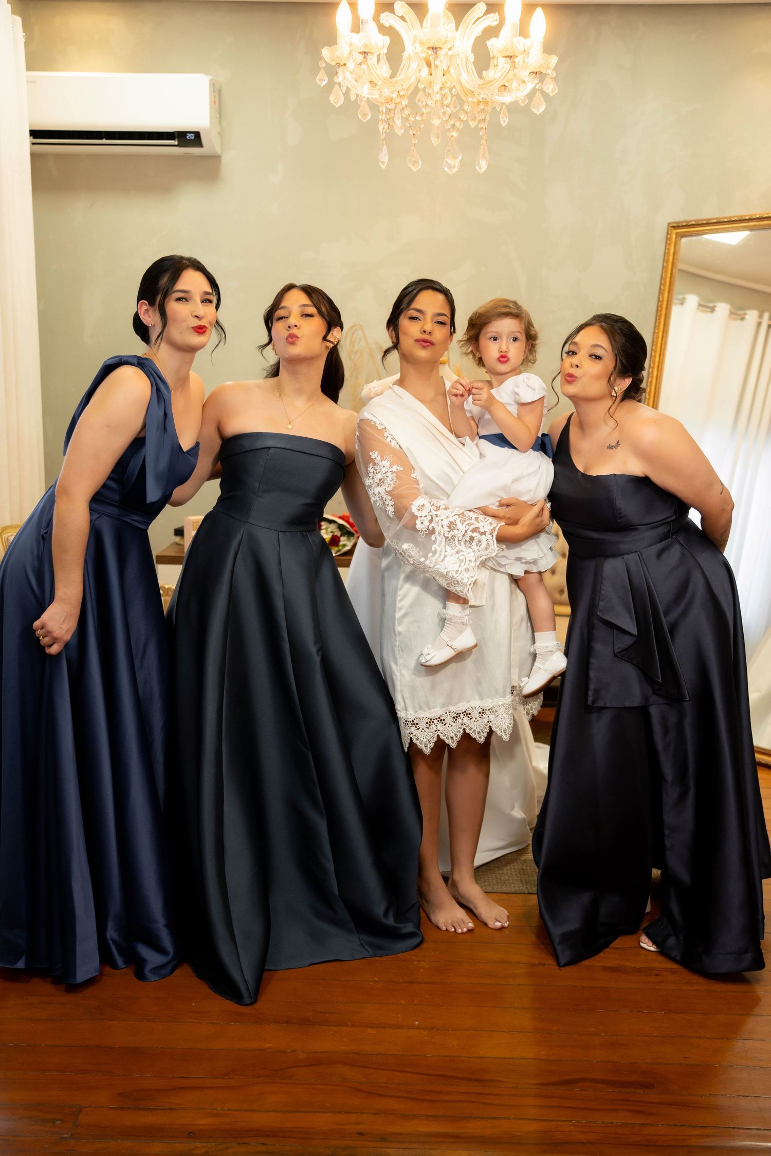 Bride in white lace robe poses with three bridesmaids in navy gowns and flower girl under chandelier.