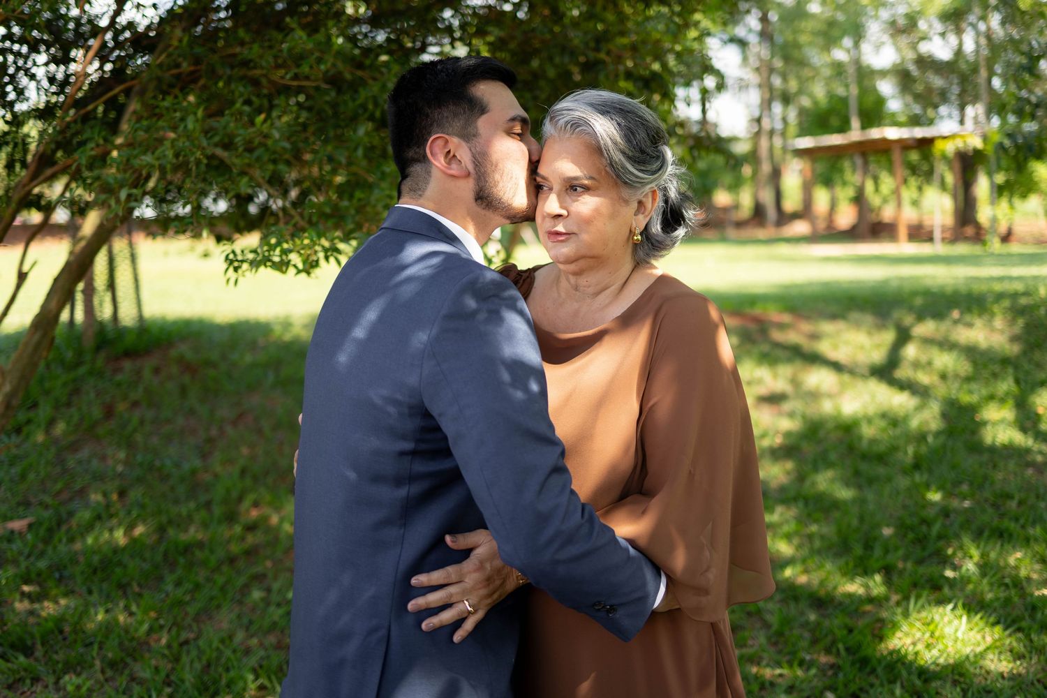 A young man in a suit kisses an older woman in a brown dress outdoors in a lush green garden.