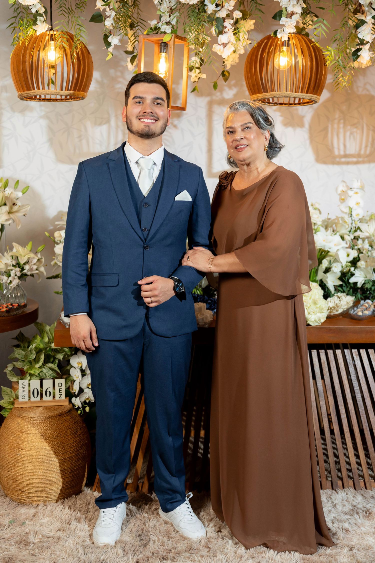 Young man in navy suit poses with older woman in brown dress at elegant floral event venue.