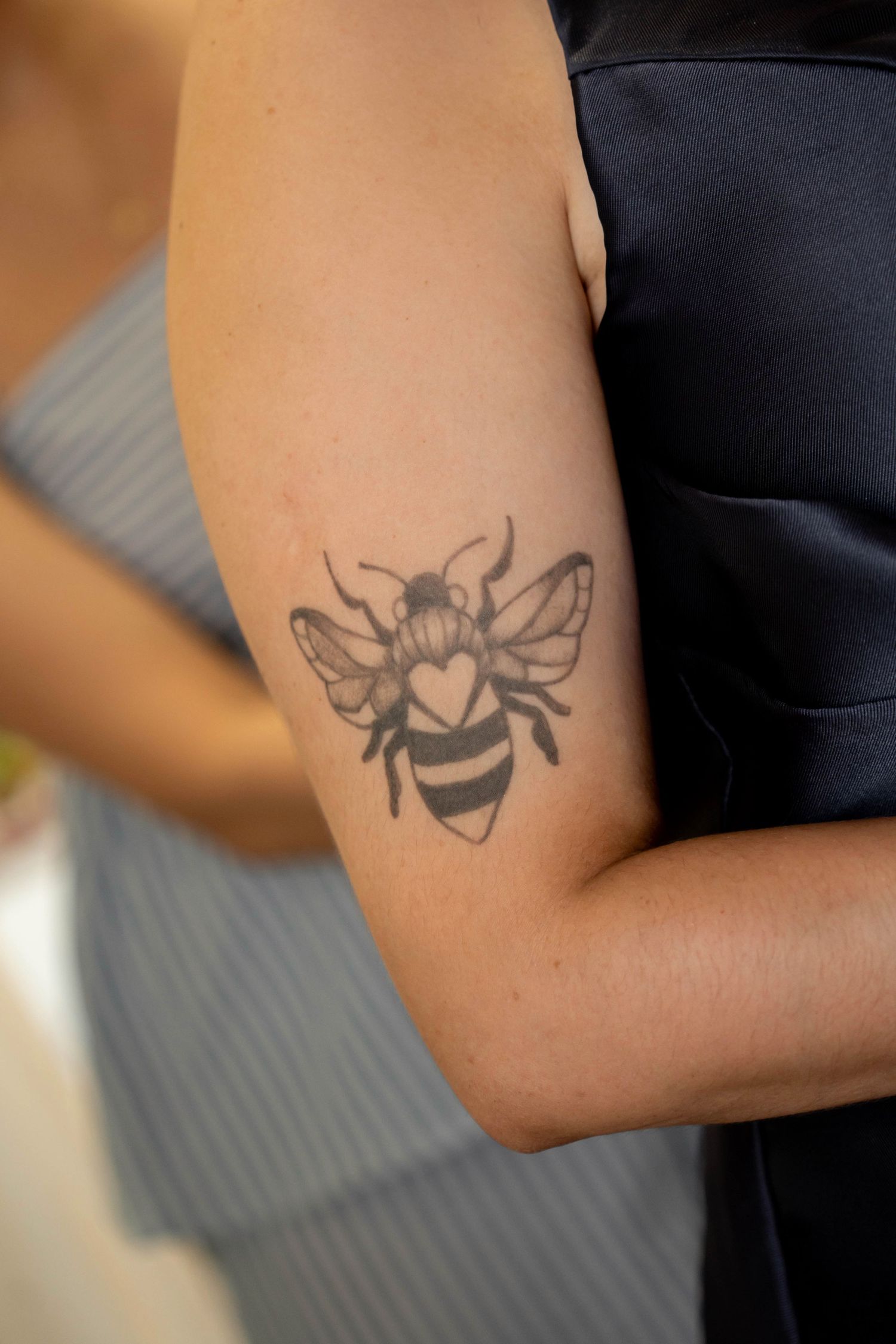 Close-up of a black and grey bee tattoo with a heart on a person's upper arm.