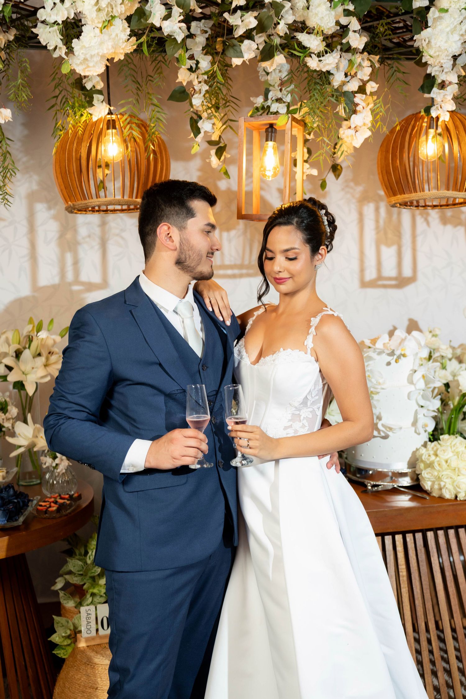 Bride and groom holding champagne glasses at elegant floral wedding reception with warm hanging lanterns.