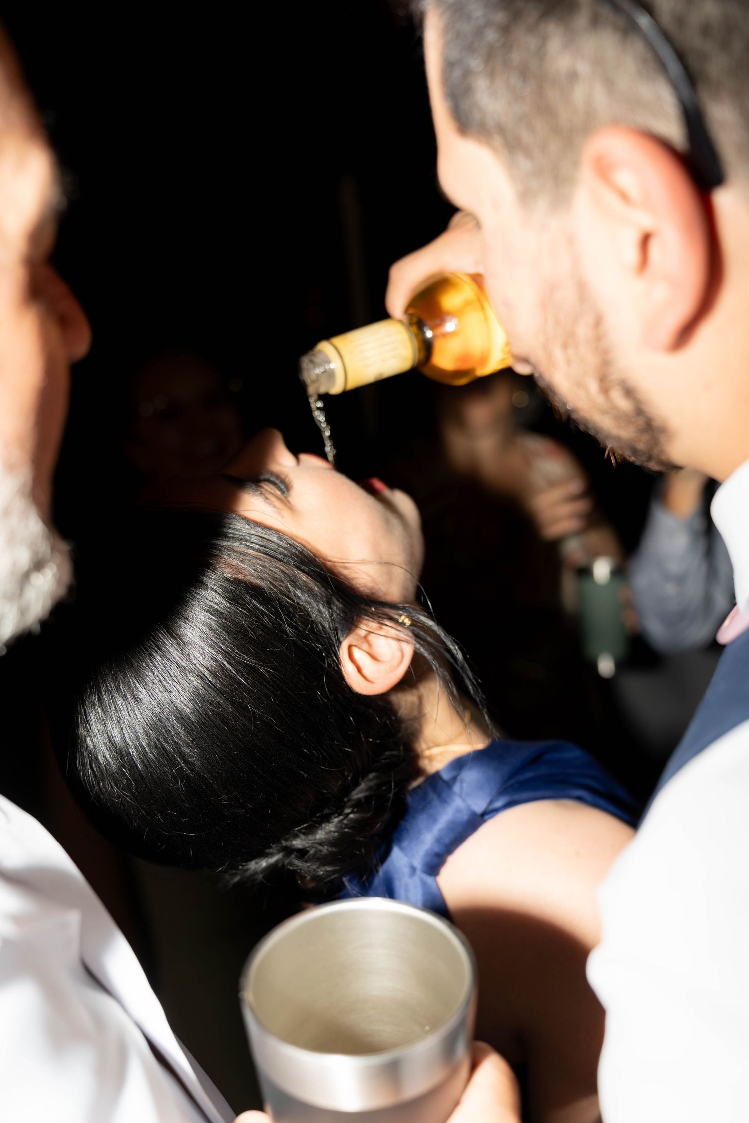 Group of people at a party, man drinking from a beer bottle while others socialize in a dimly lit venue.