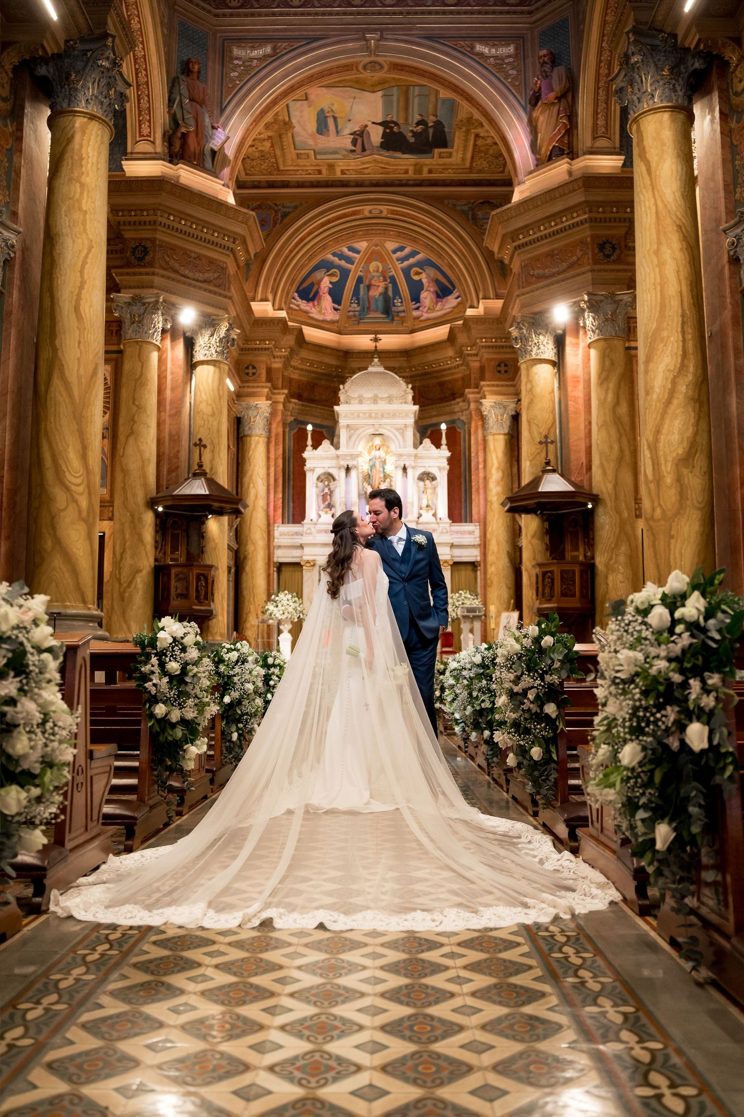 Bride and groom share a kiss at the altar of an ornate church, with floral arrangements and a long bridal veil.