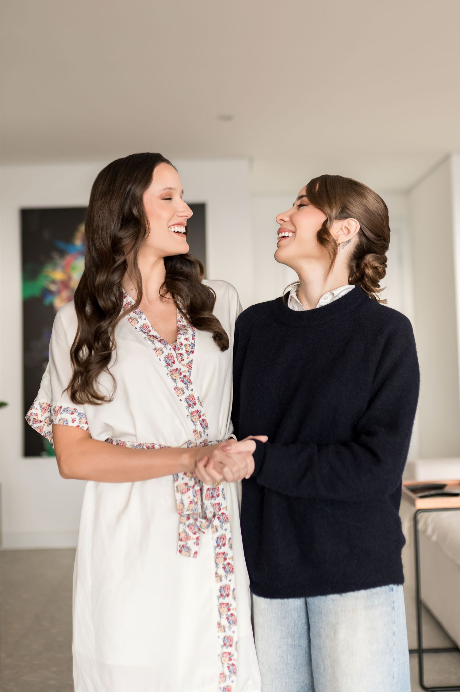 Two women smiling and holding hands indoors, one in a floral robe and one in a navy sweater with jeans.