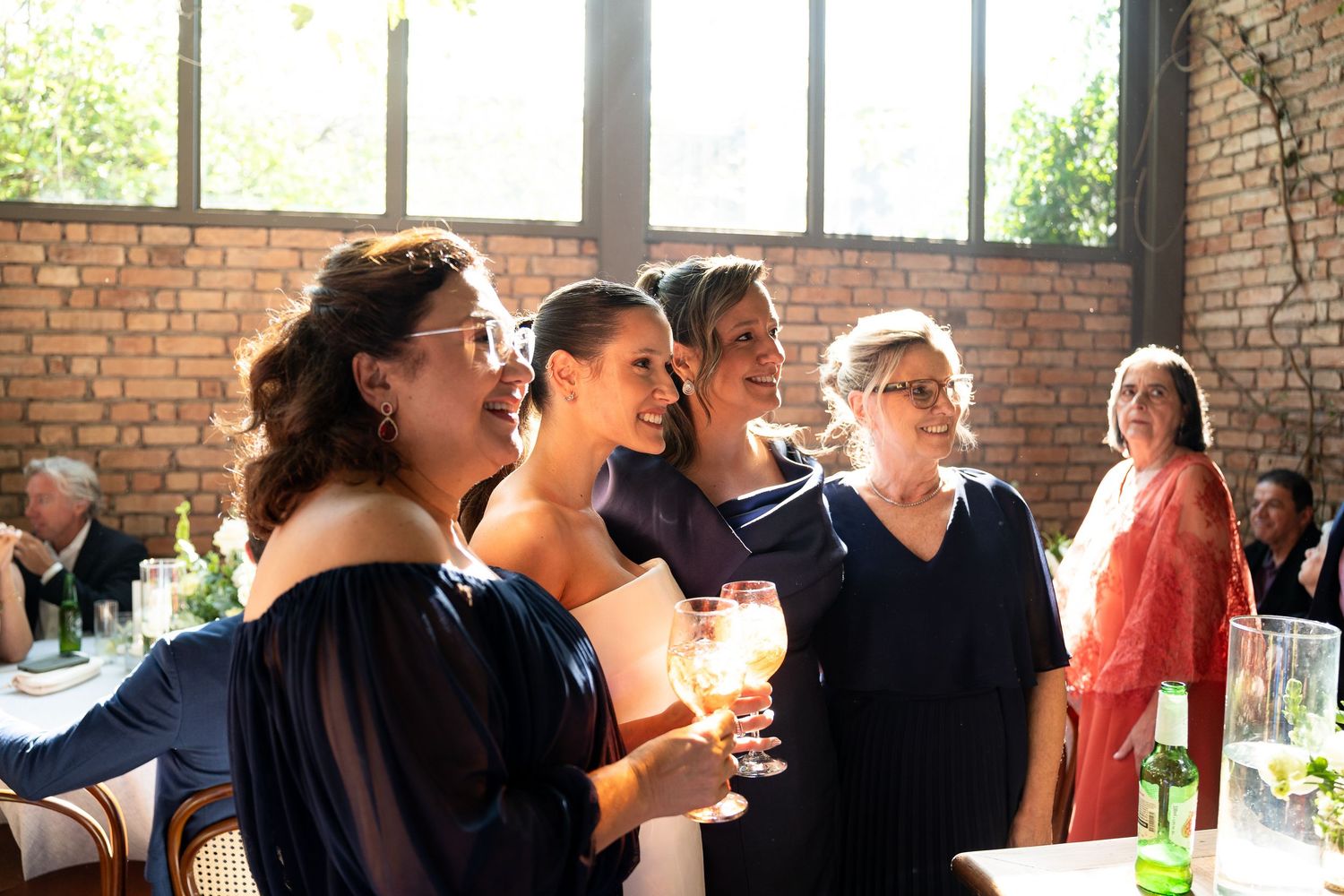 Women in elegant dresses smiling and holding drinks at a wedding reception in a brick-walled venue.