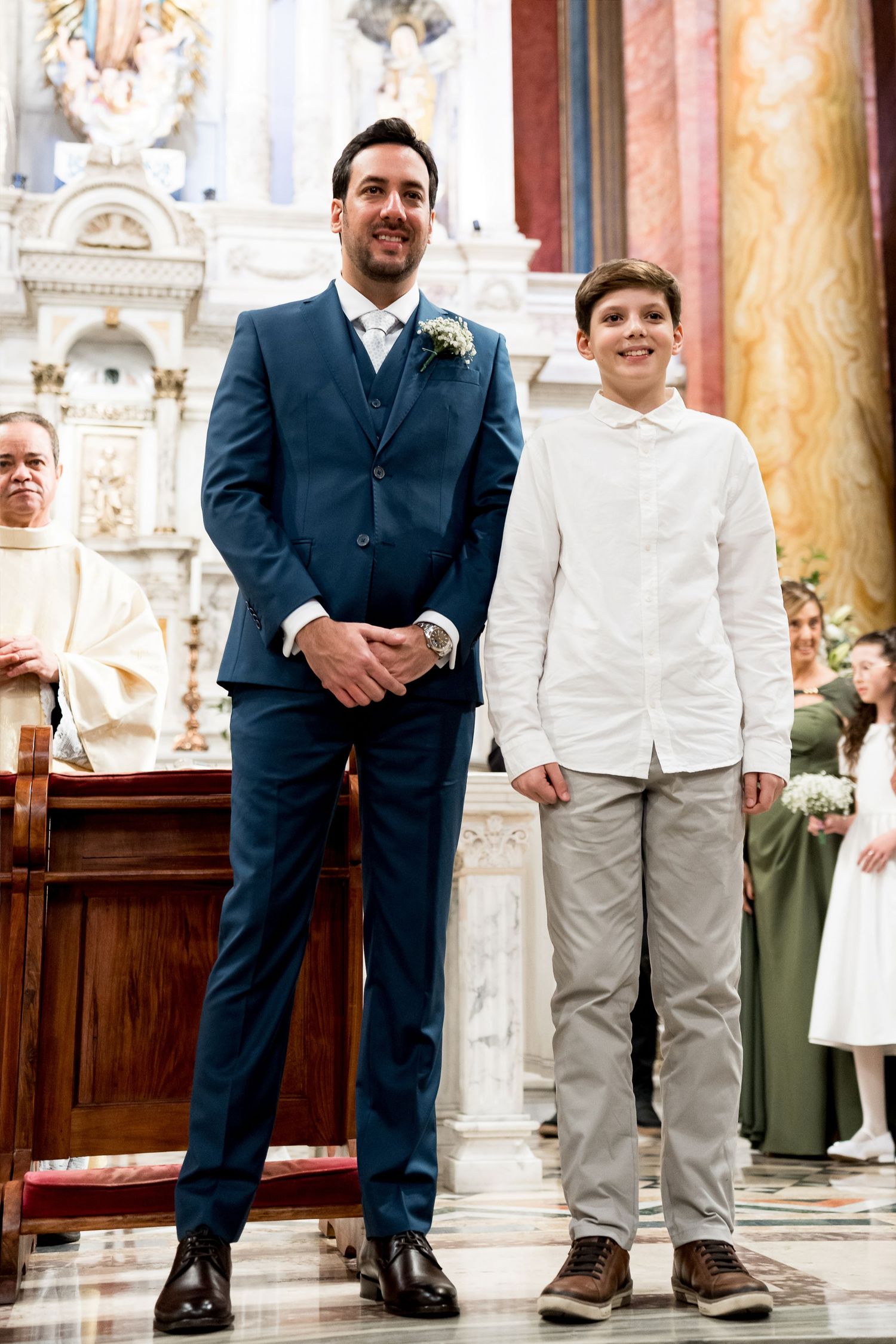 Man in blue suit and boy in white shirt stand smiling together inside an ornate church during a formal ceremony.