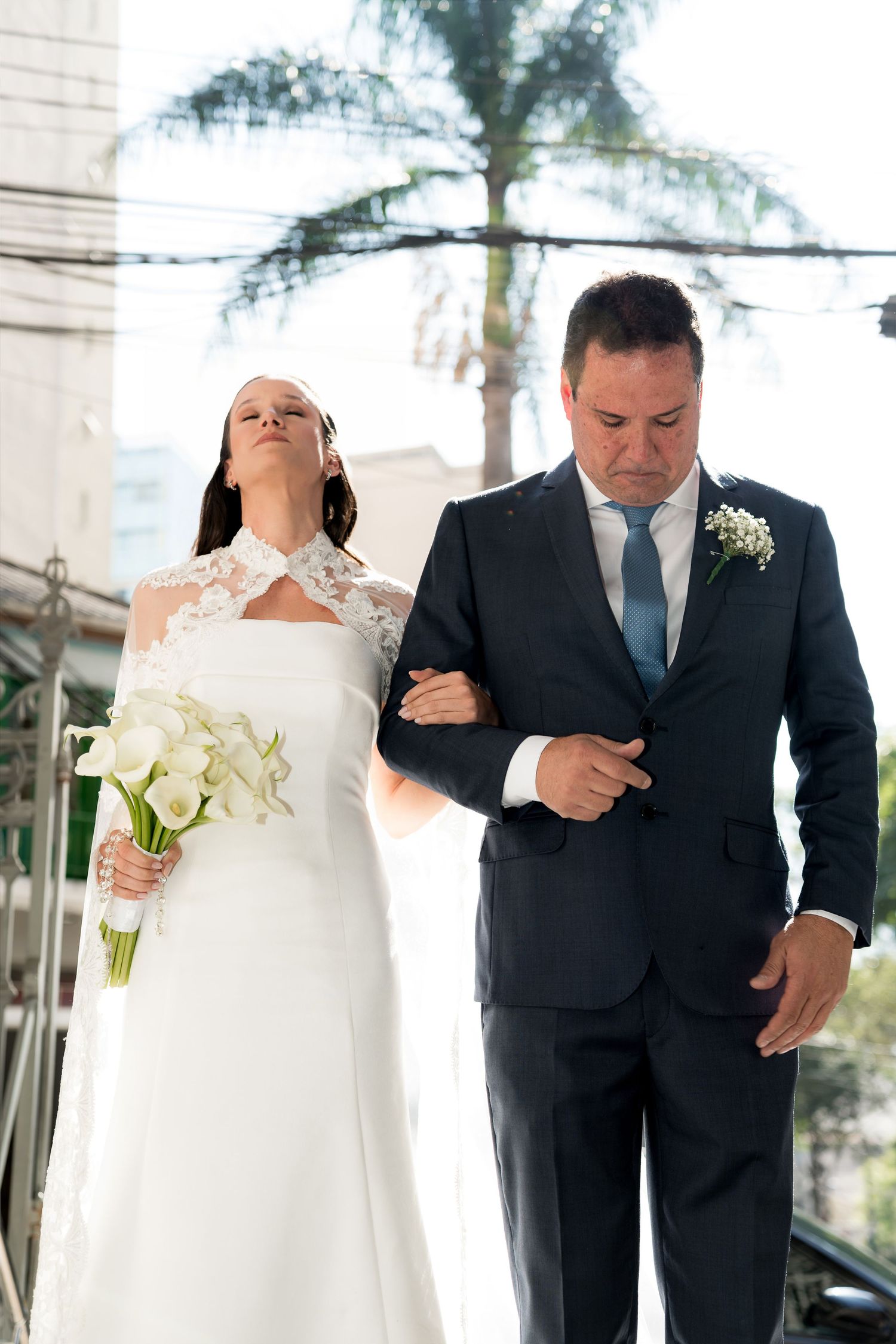 Bride in white lace gown holding calla lily bouquet walks arm-in-arm with groom in dark suit and blue tie.