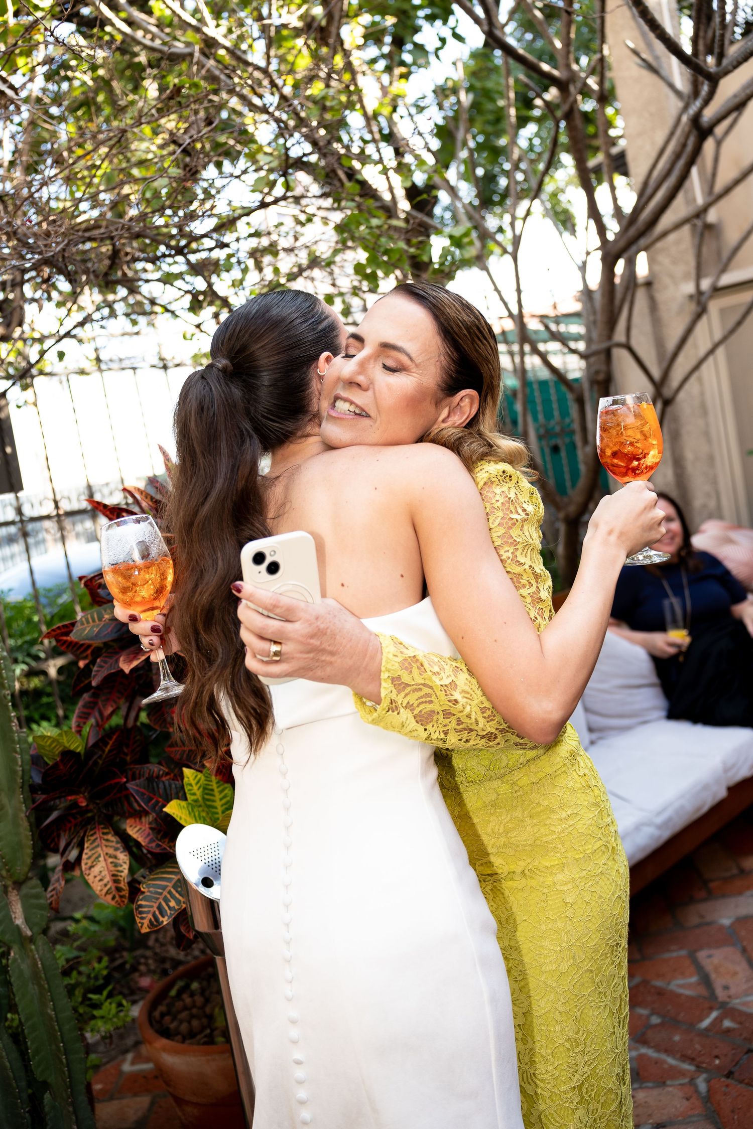 Two women embracing at a wedding, one in a white gown, the other in yellow lace, holding orange cocktails outdoors.