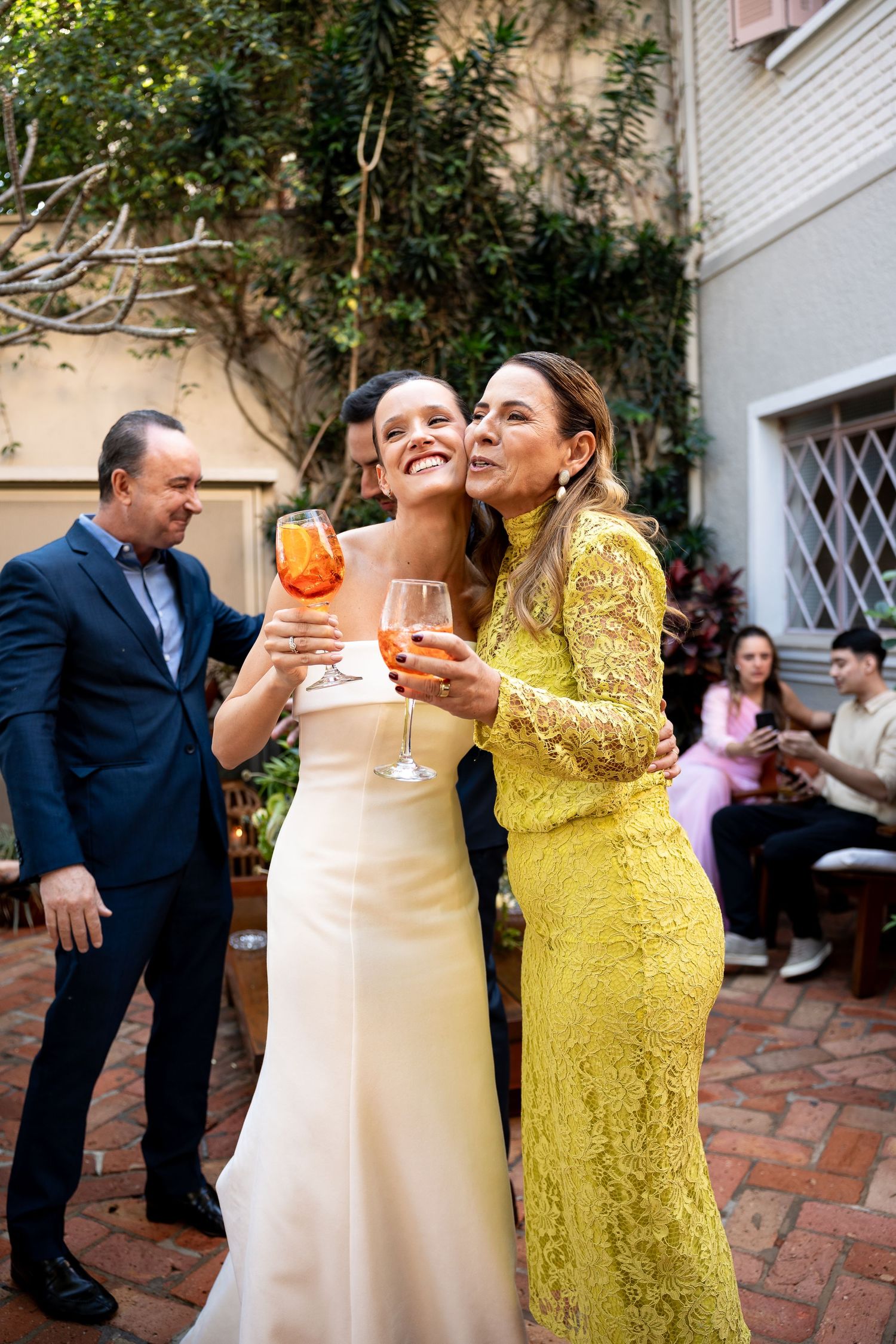 Bride in white gown and woman in yellow lace dress toast with Aperol Spritz at an outdoor wedding celebration.