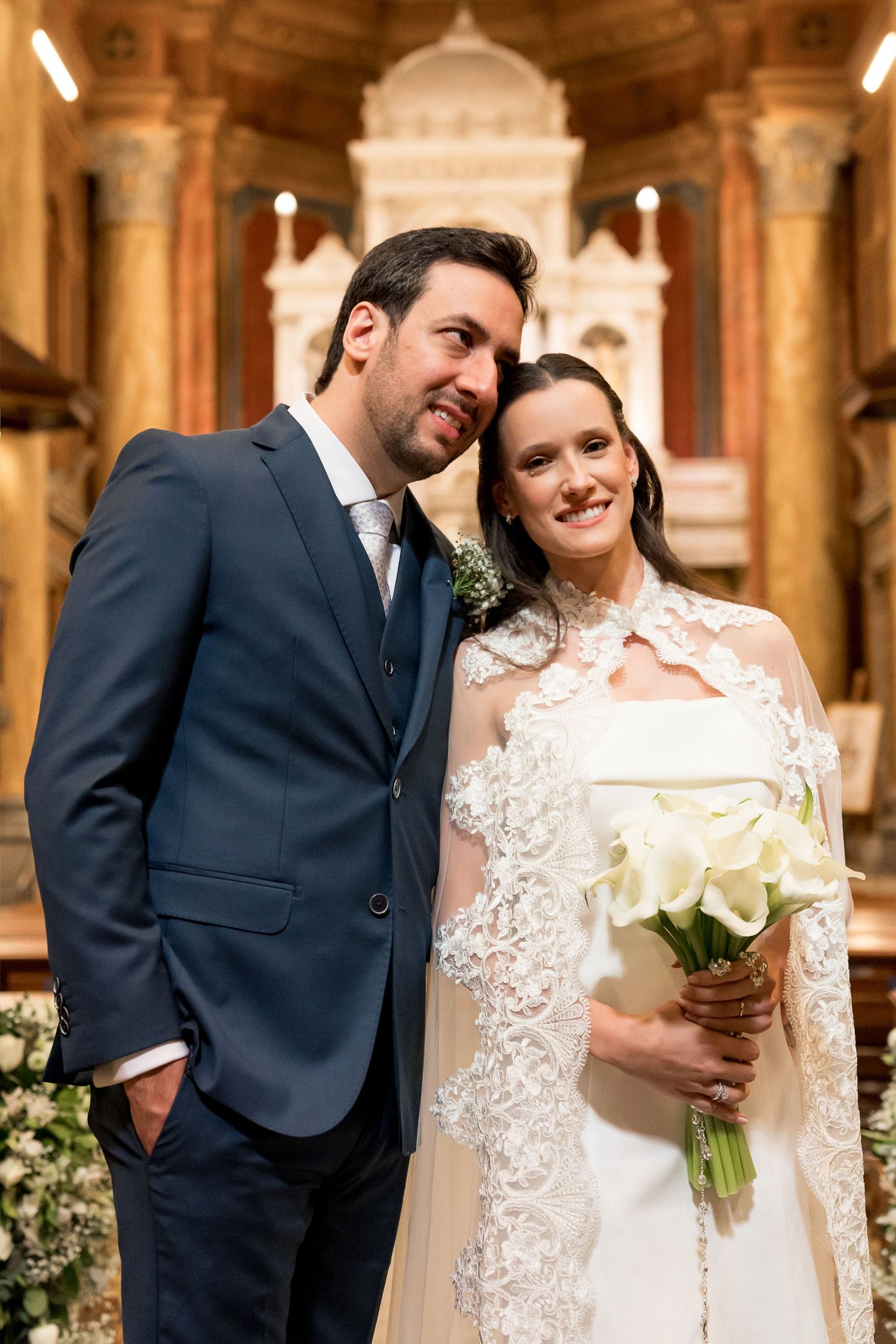 Elegant bride and groom posing together inside a church, bride holding white calla lily bouquet in lace wedding gown.
