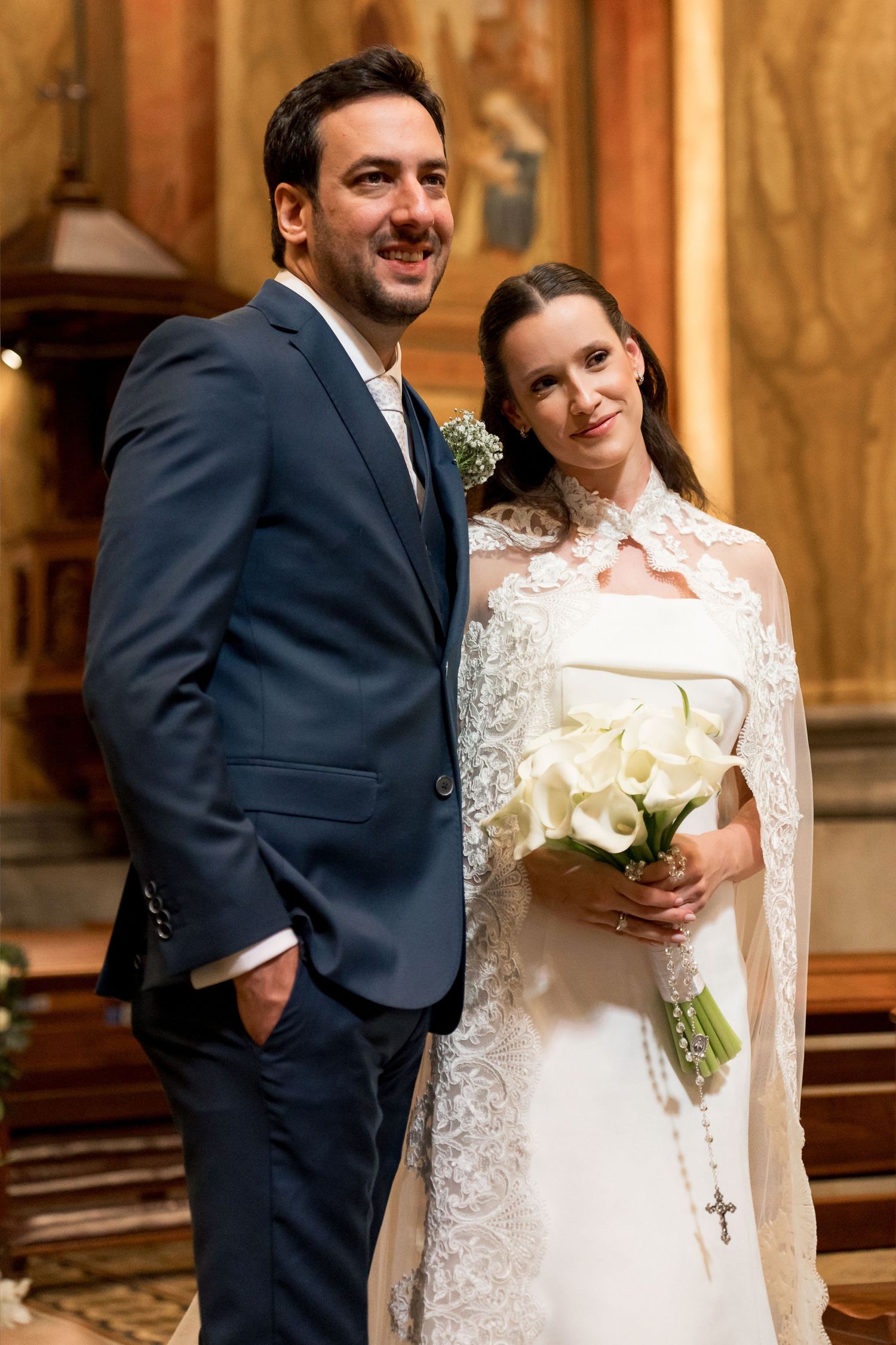 Bride and groom posing together inside a church, bride holding white calla lily bouquet wearing lace wedding gown.