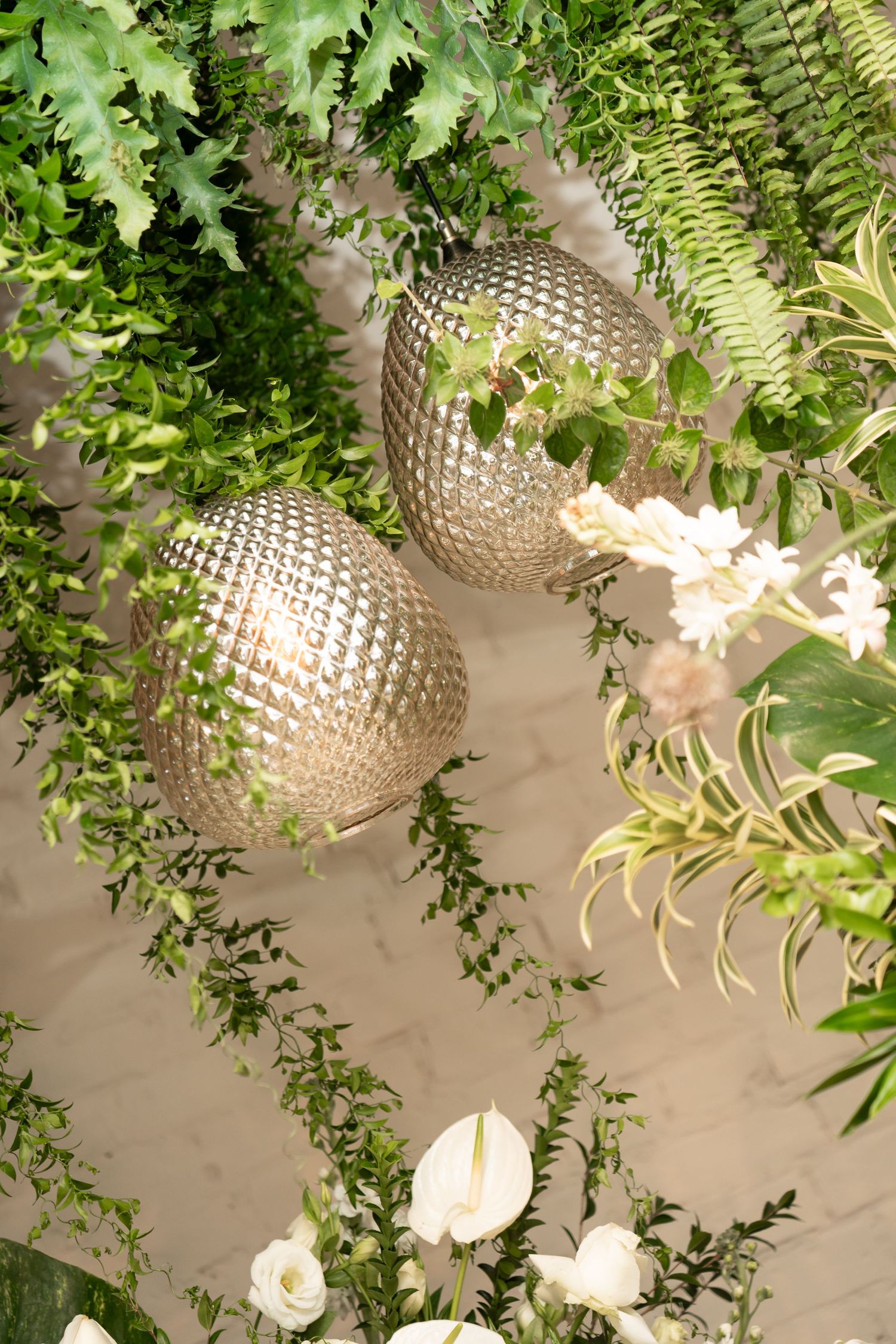 Gold geometric pendant lights hanging among lush green ferns and white flowers in elegant floral installation.