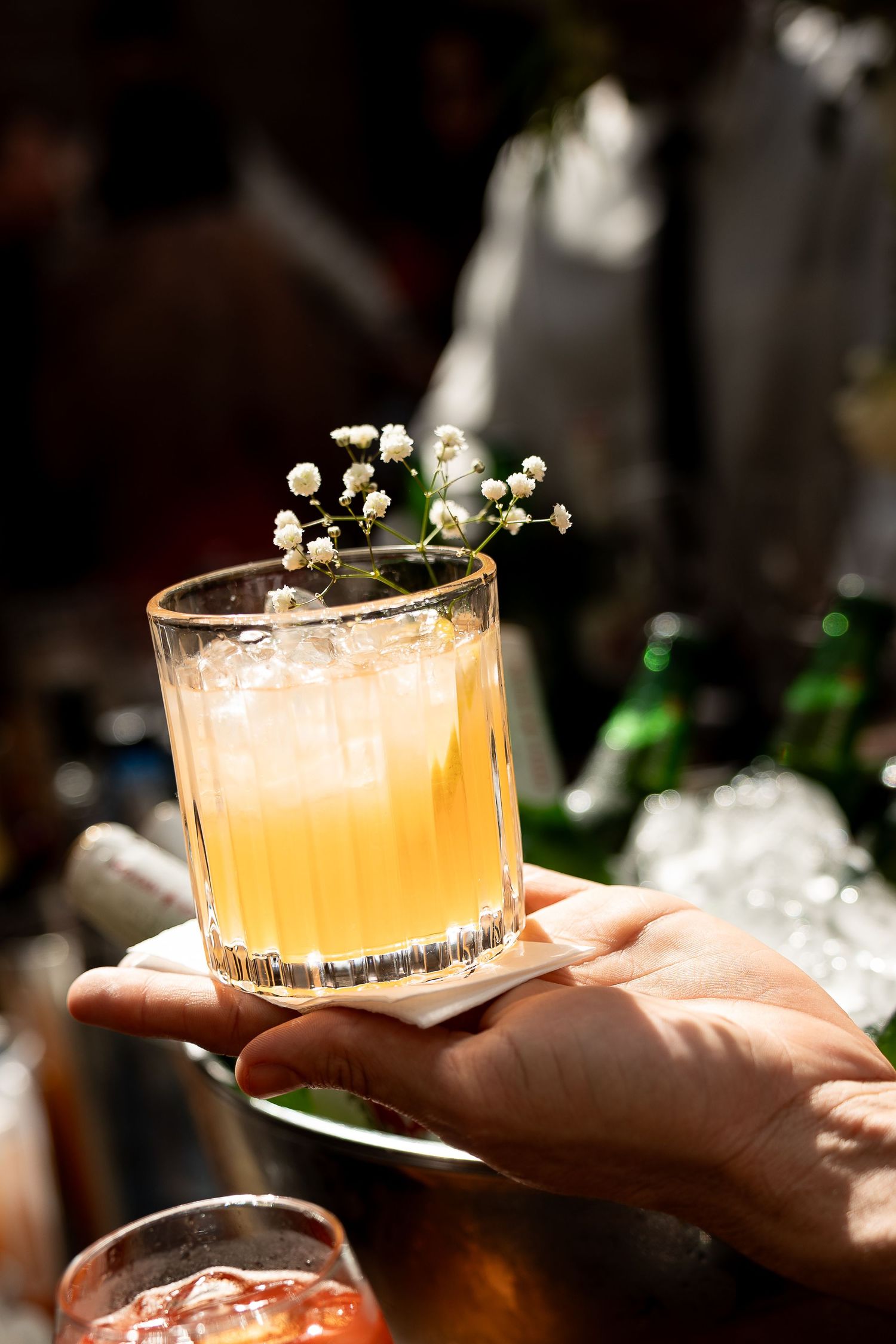 Hand holding a rocks glass with golden cocktail, ice, and delicate white flowers garnish at a bar.
