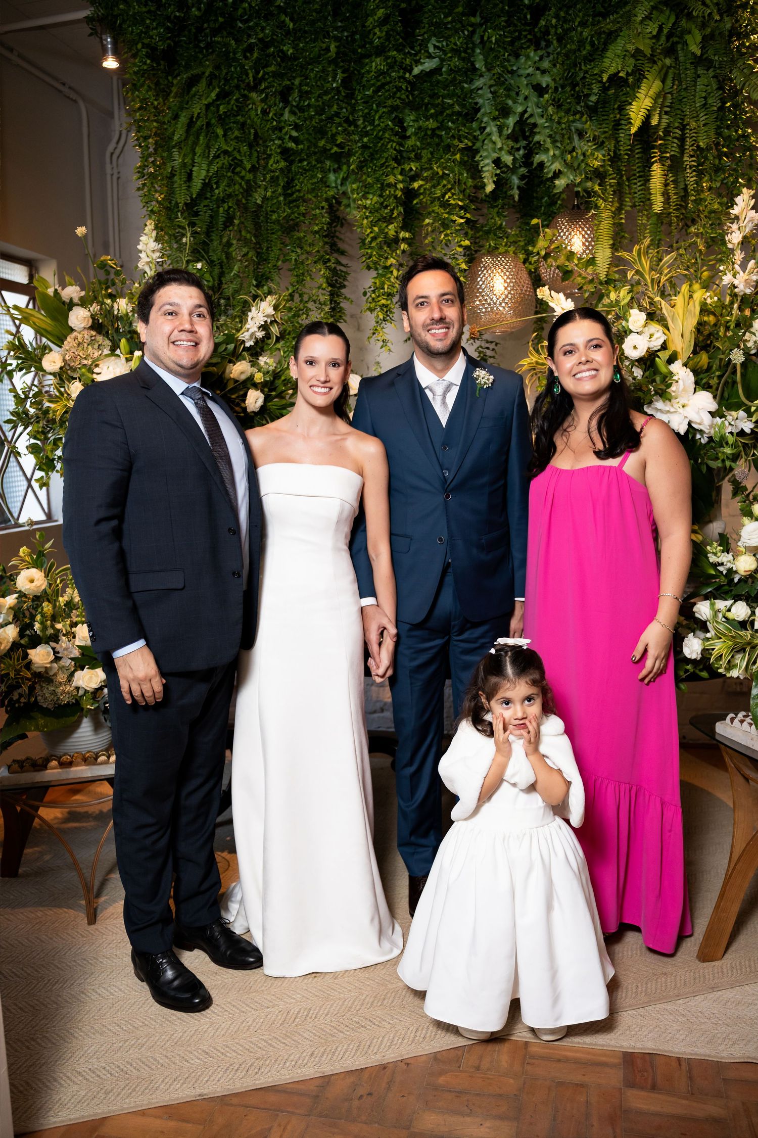 Wedding party poses with bride in white gown, groom in suit, guests in pink dress and navy suit, and flower girl.