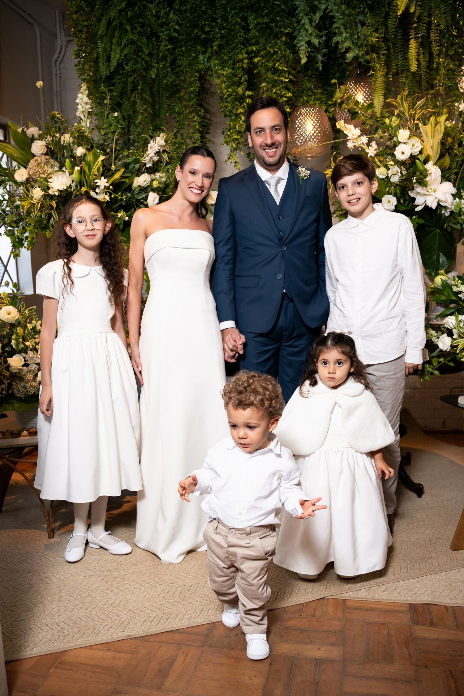 A bride and groom pose with four children in white attire before a lush floral backdrop at an elegant wedding ceremony.