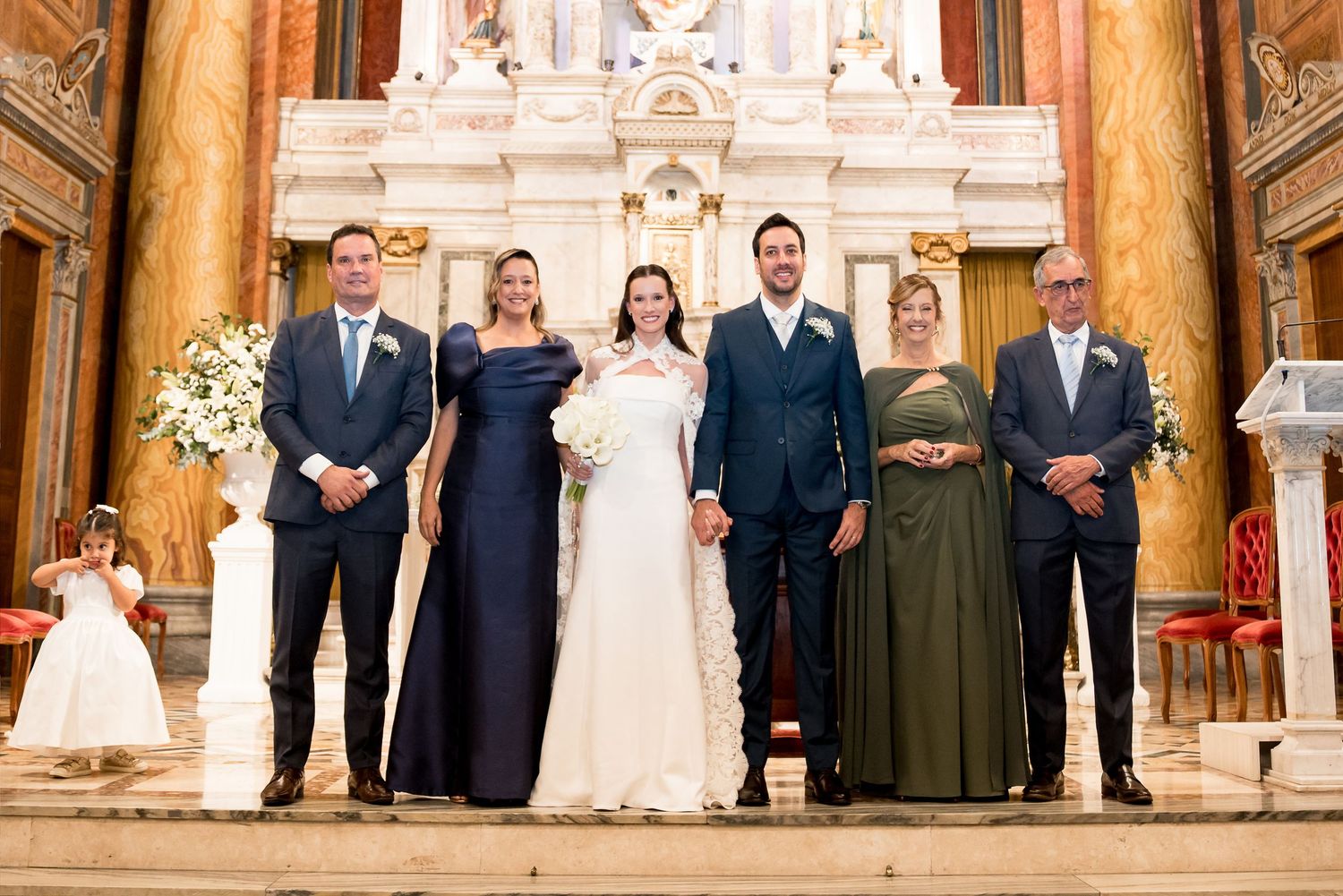 Wedding party posing in an ornate church, bride in white gown with groom and family members in formal attire.