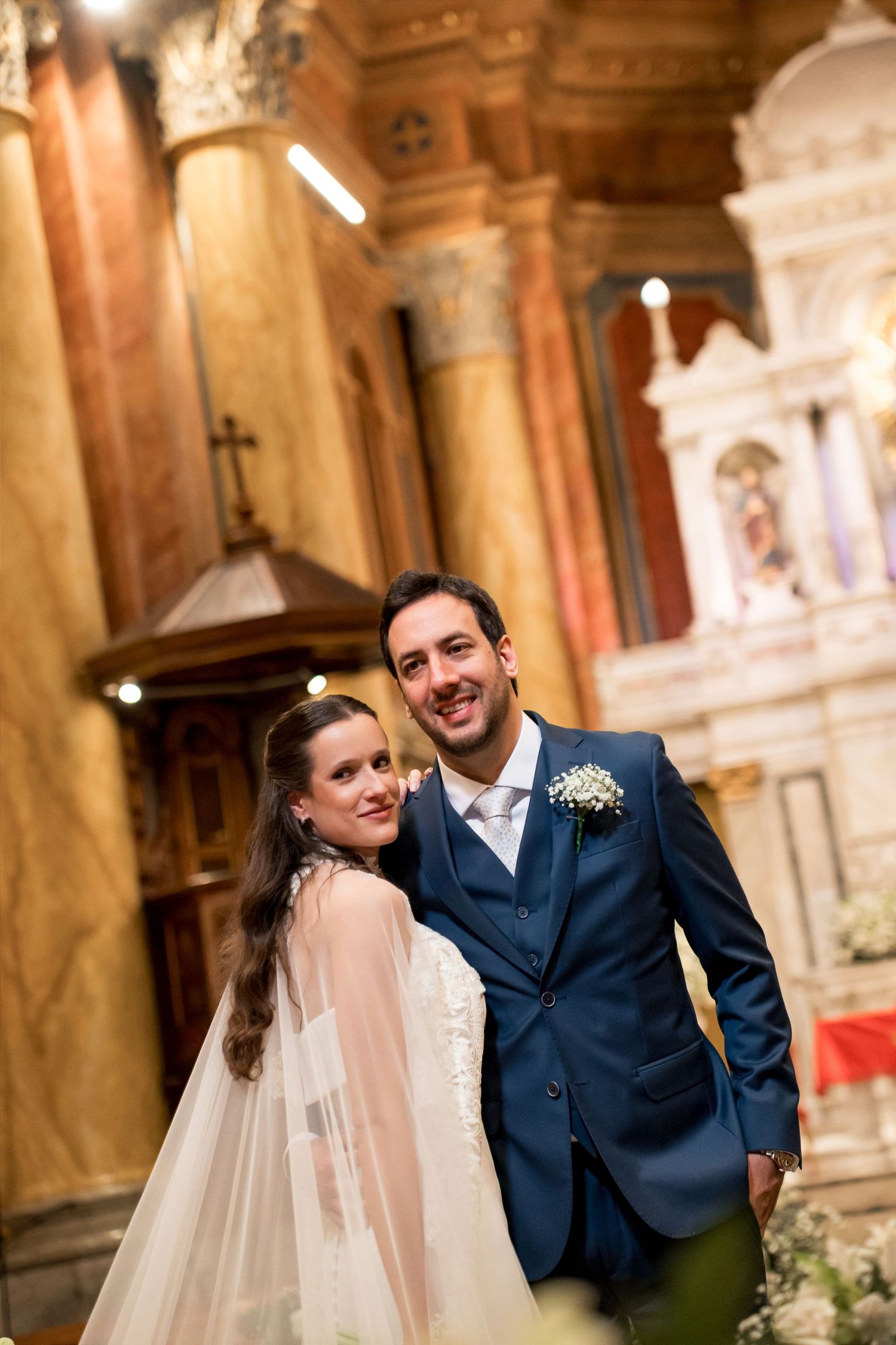 Bride and groom posing together inside an ornate church, smiling, with marble columns and altar in the background.