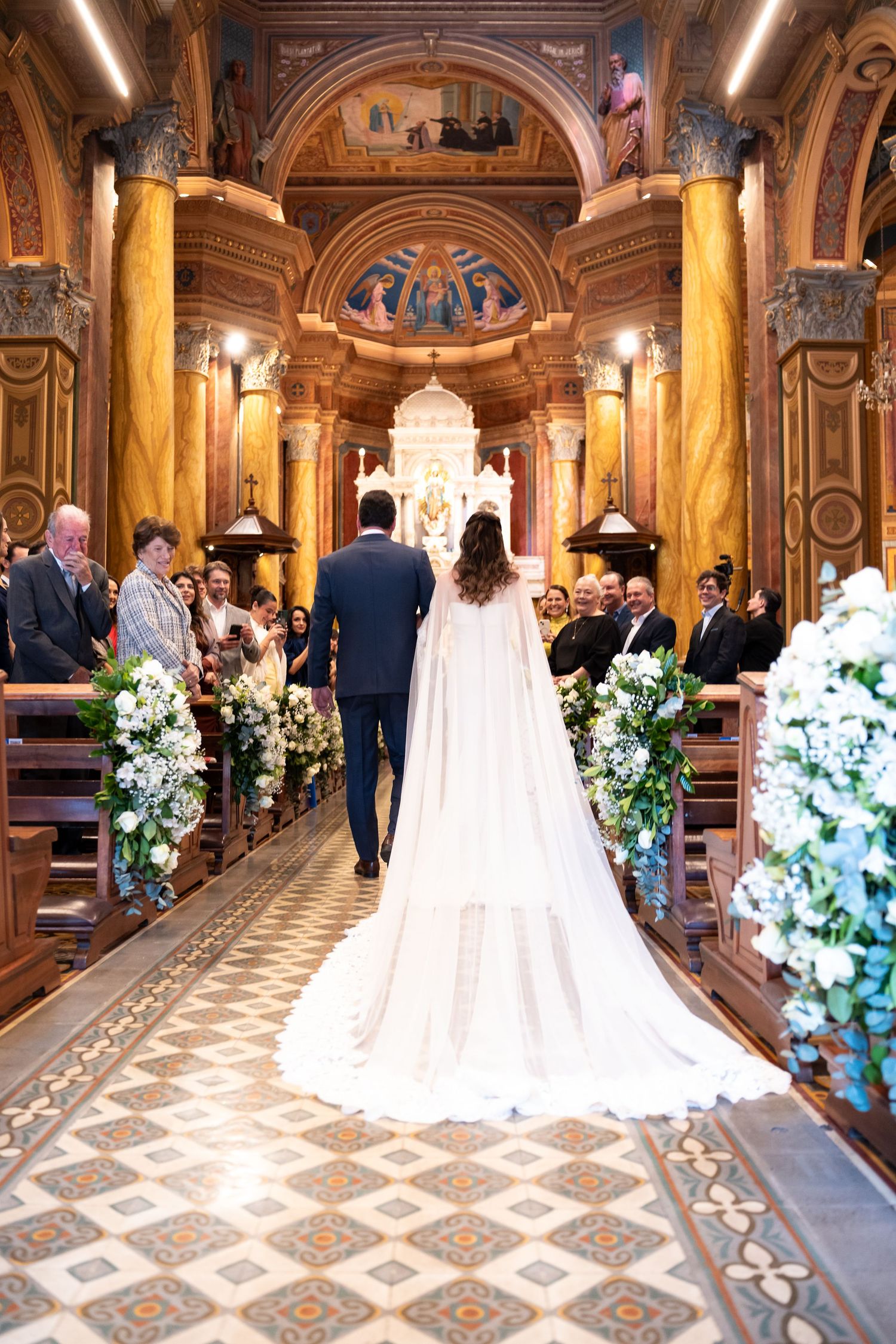 Bride and groom walking down the aisle of an ornate church decorated with white floral arrangements.