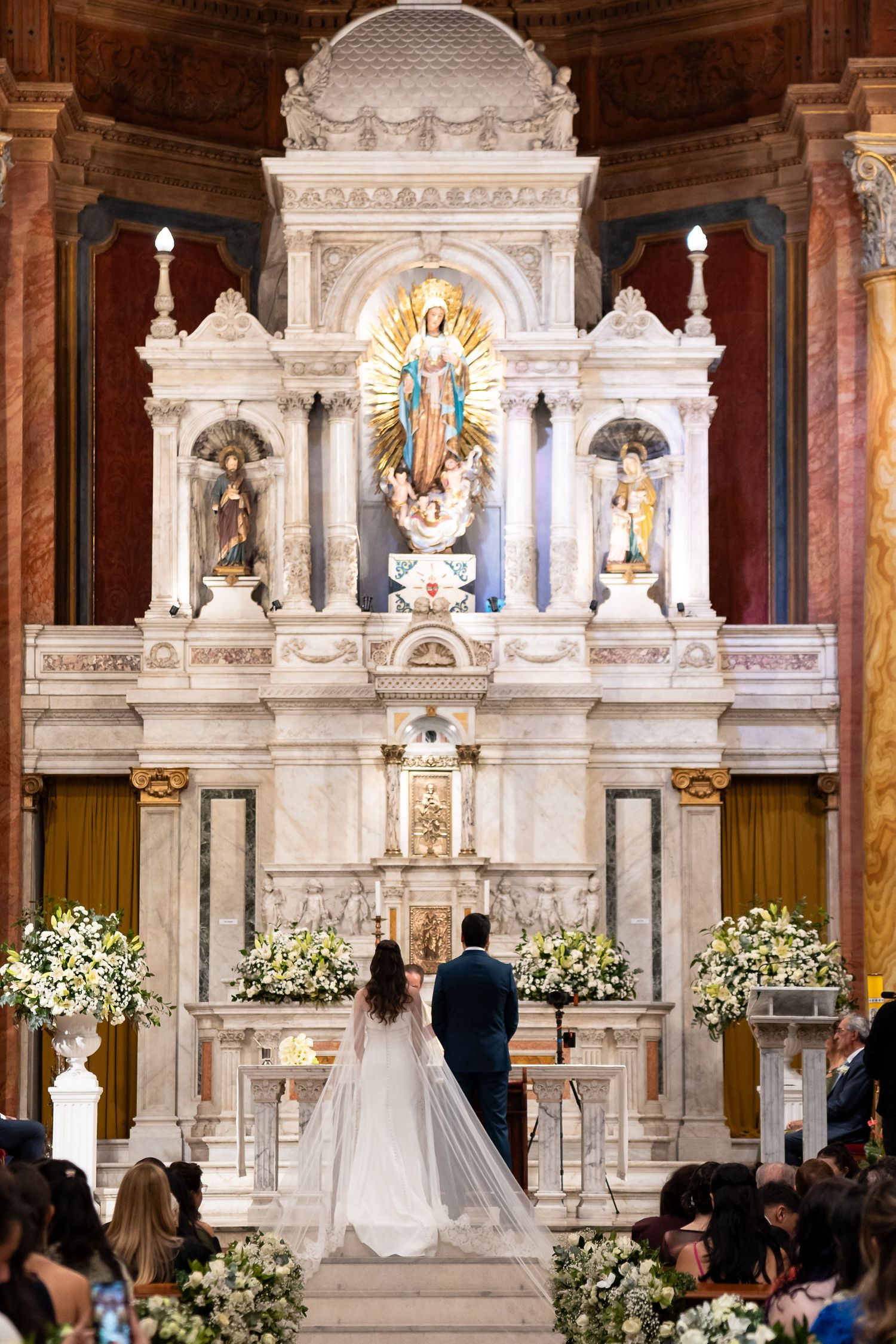 Bride and groom stand before ornate white marble altar with floral arrangements in elegant Catholic church wedding ceremony.