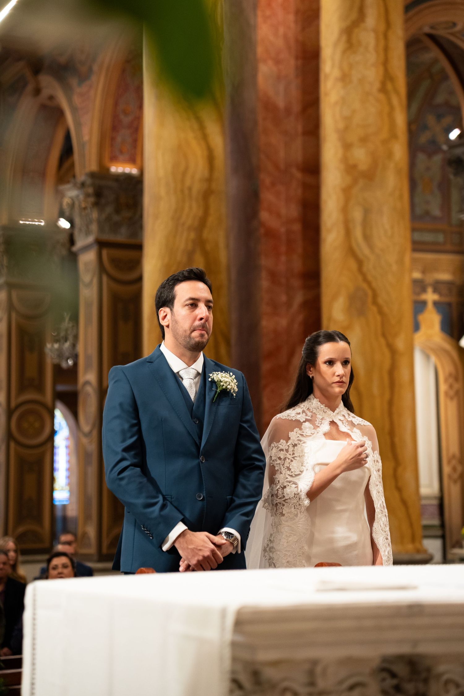 Bride in lace cape and groom in blue suit stand at church altar during elegant wedding ceremony.